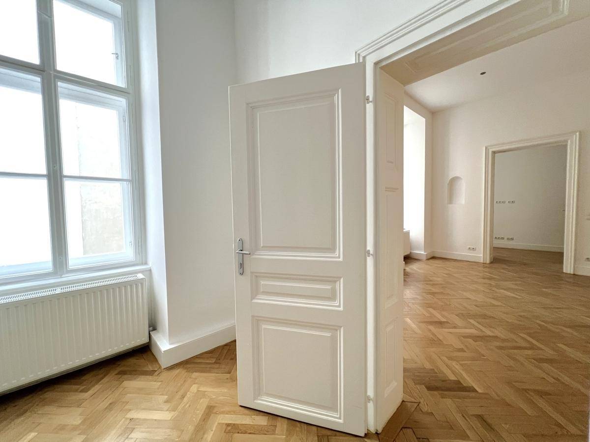 Offene weiße Tür in einem hellen Zimmer mit klassischem Fischgrätparkett und hohen Fenstern.