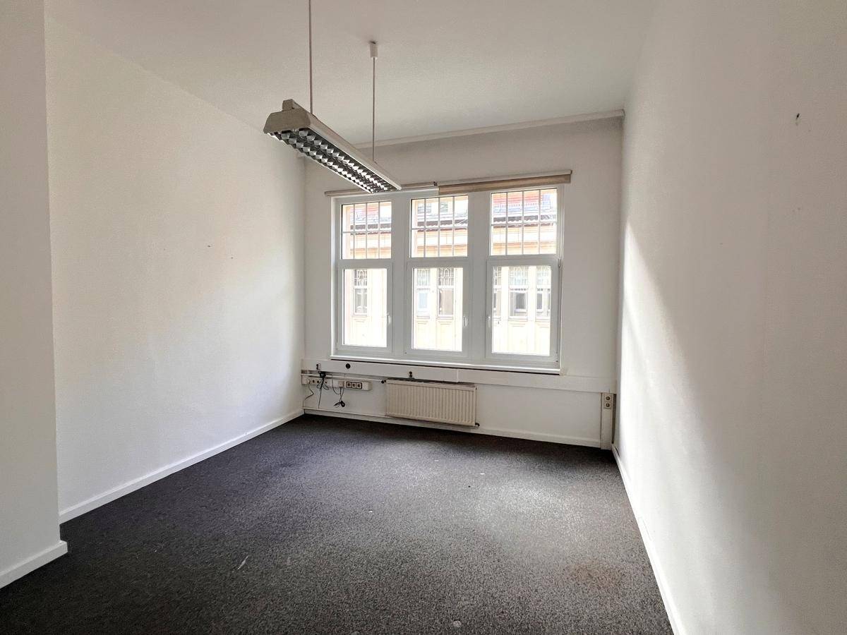 Leeres Büro mit dunklem Teppichboden, weißen Wänden und einem großen Fenster, das für ausreichend Helligkeit sorgt.