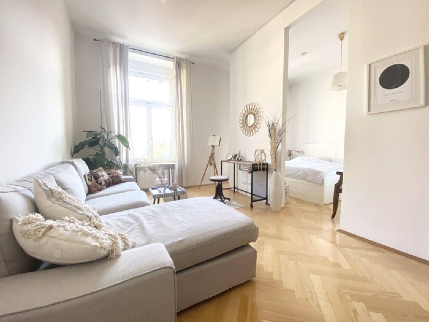 Heller Wohnbereich mit bequemem Sofa und offenem Übergang zum Schlafzimmer, ausgestattet mit Fischgrätparkett.