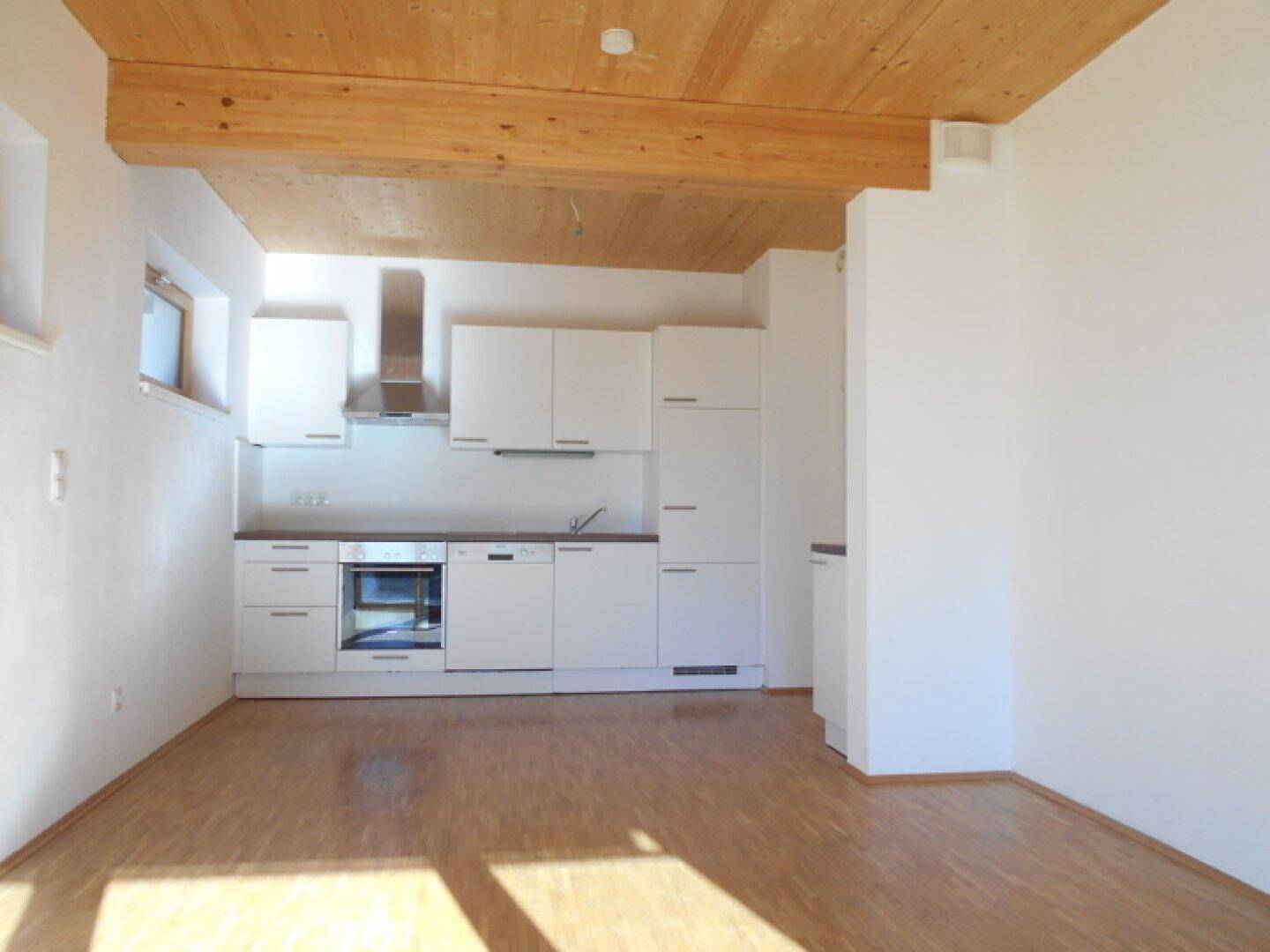 Moderne Einbauküche mit weißen Schränken, integrierten Geräten und Holzbalkendecke in einem hellen Raum.
