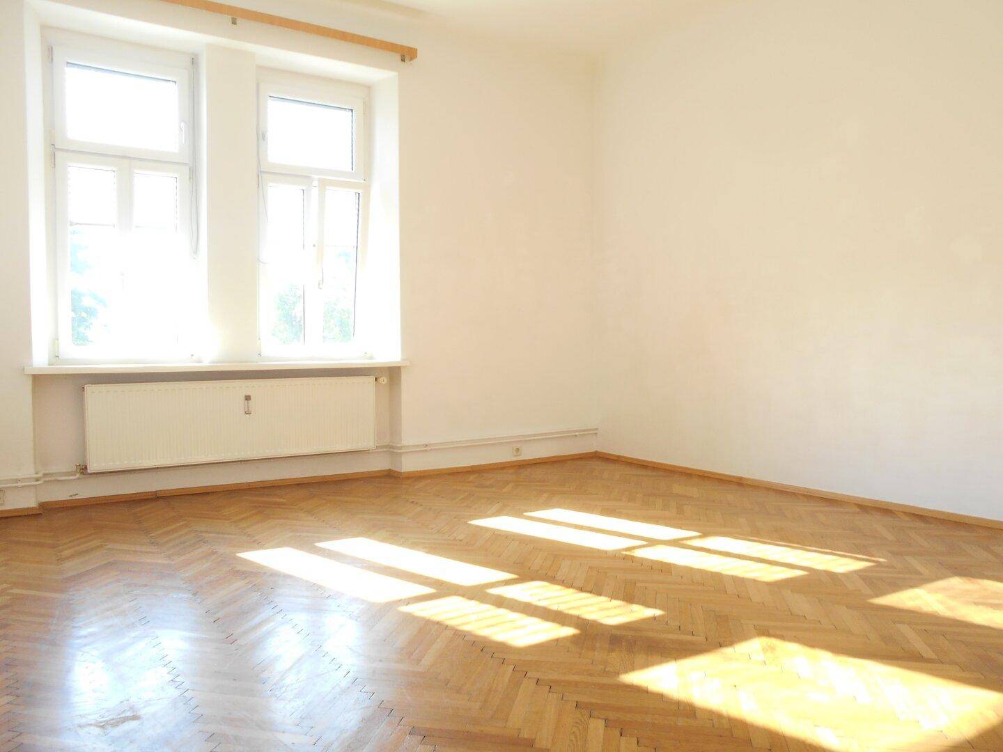Heller Raum mit Fischgrätparkett, weißen Wänden und einem großen Fenster, das viel Sonnenlicht hereinlässt.