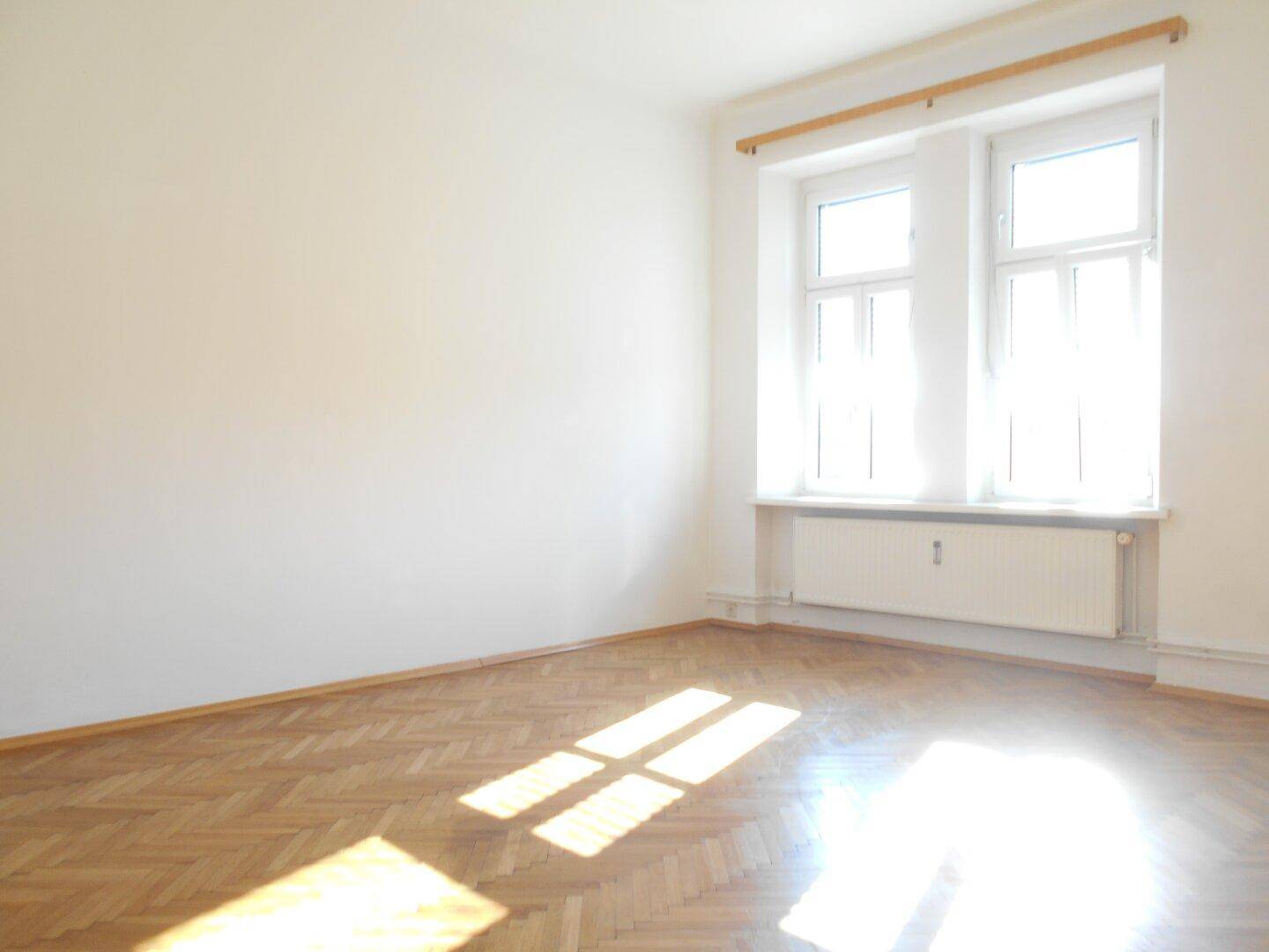 Geräumiger, heller Raum mit Fischgrätparkett und einem großen Fenster, das den Boden mit Sonnenlicht erhellt.
