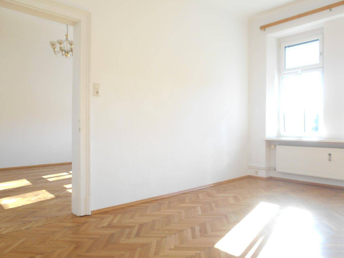 Heller Raum mit Fischgrätparkett und einem Fenster, das viel Tageslicht hereinlässt, mit offenem Durchgang.