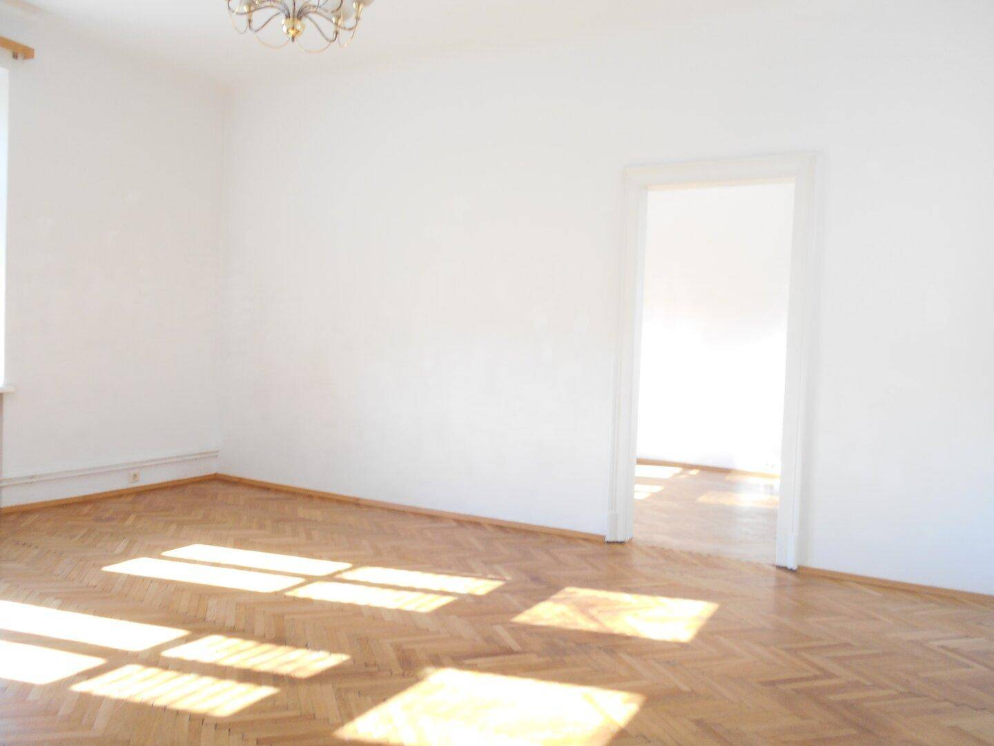 Lichtdurchfluteter Raum mit Fischgrätparkett und offenem Durchgang zu einem weiteren hellen Zimmer.