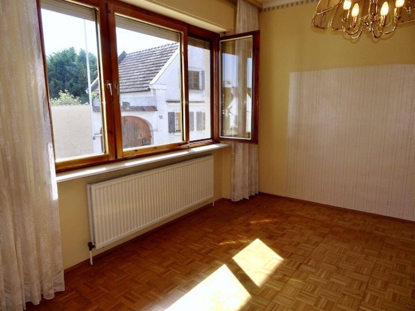 Helles Zimmer mit Fischgrätparkettboden, großen Fenstern und einem Heizkörper unter der Fensterbank.