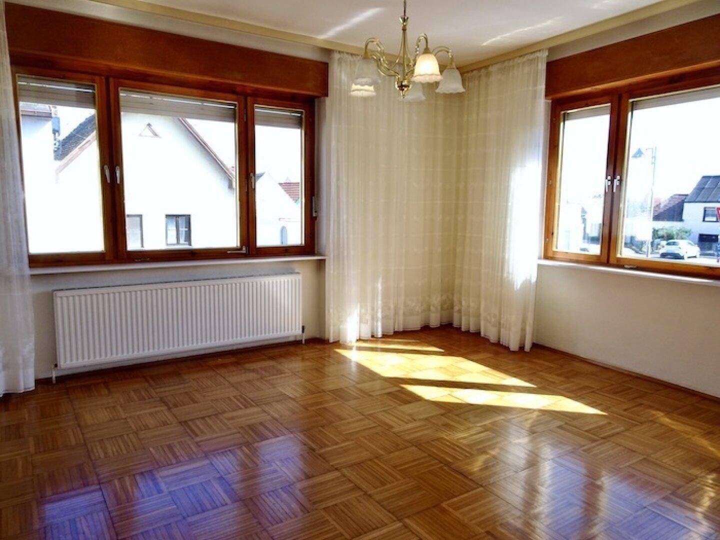 Lichtdurchfluteter Raum mit klassischem Parkettboden, großen Fenstern und einem Kronleuchter an der Decke.