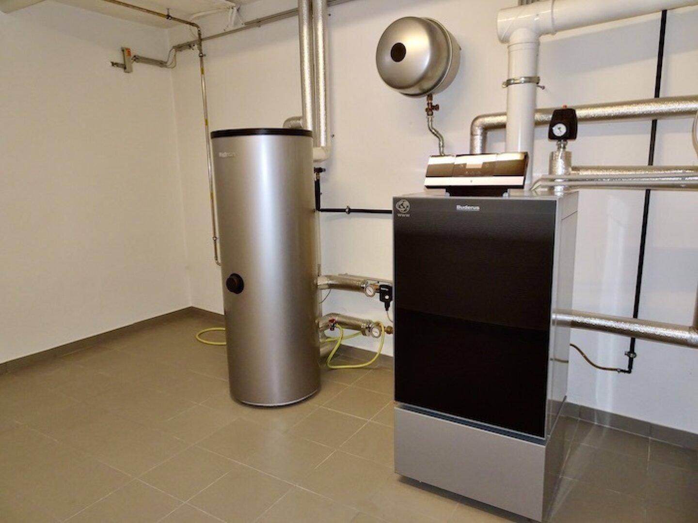 Heizungsraum mit moderner Heizungsanlage und Warmwasserspeicher auf gefliestem Boden.