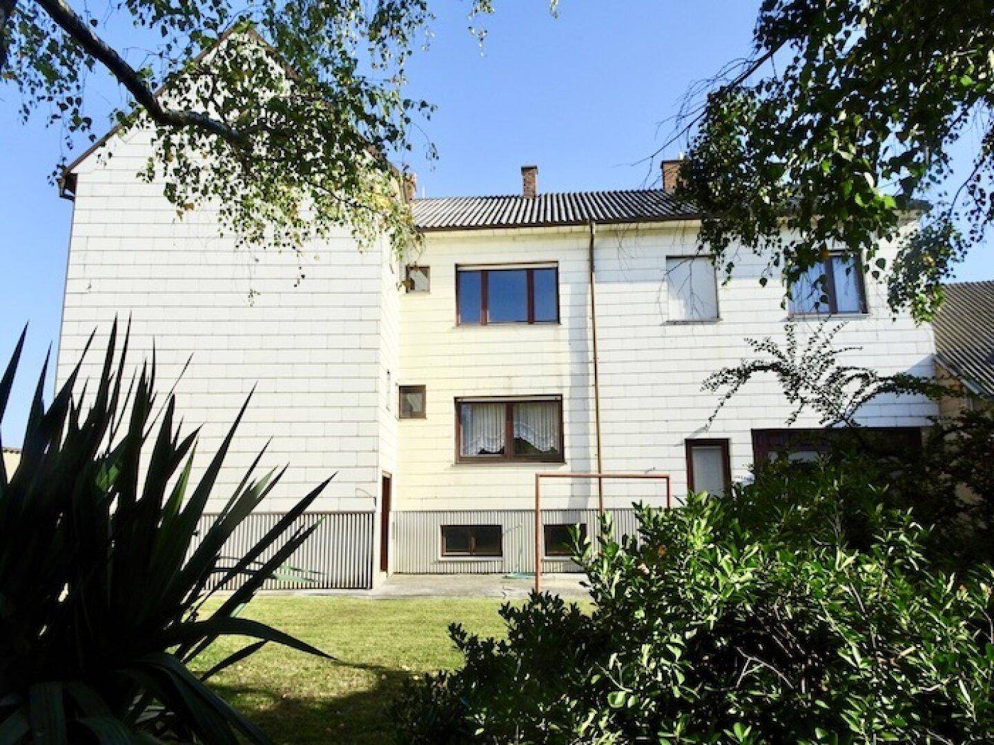 Rückseite des Hauses mit weißer Fassade, Fenstern und einem kleinen Gartenbereich mit Rasen.