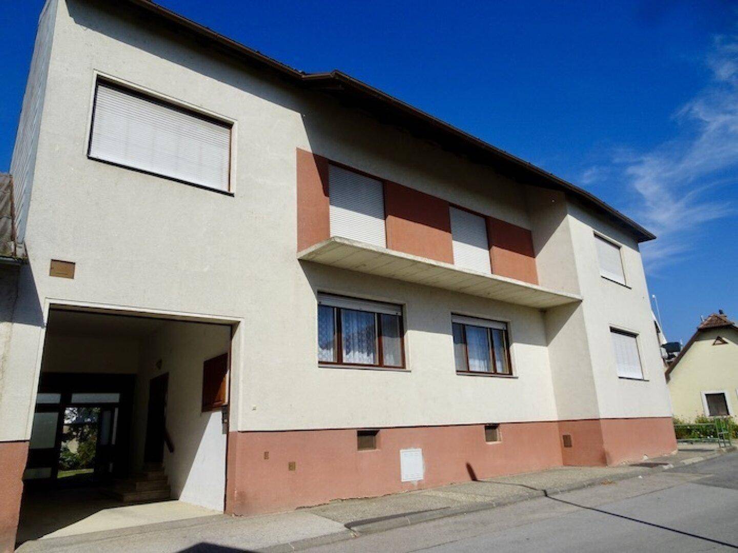 Ansicht des Gebäudes von der Straße mit Eingangsbereich und Fenstern mit Rollläden bei sonnigem Wetter.