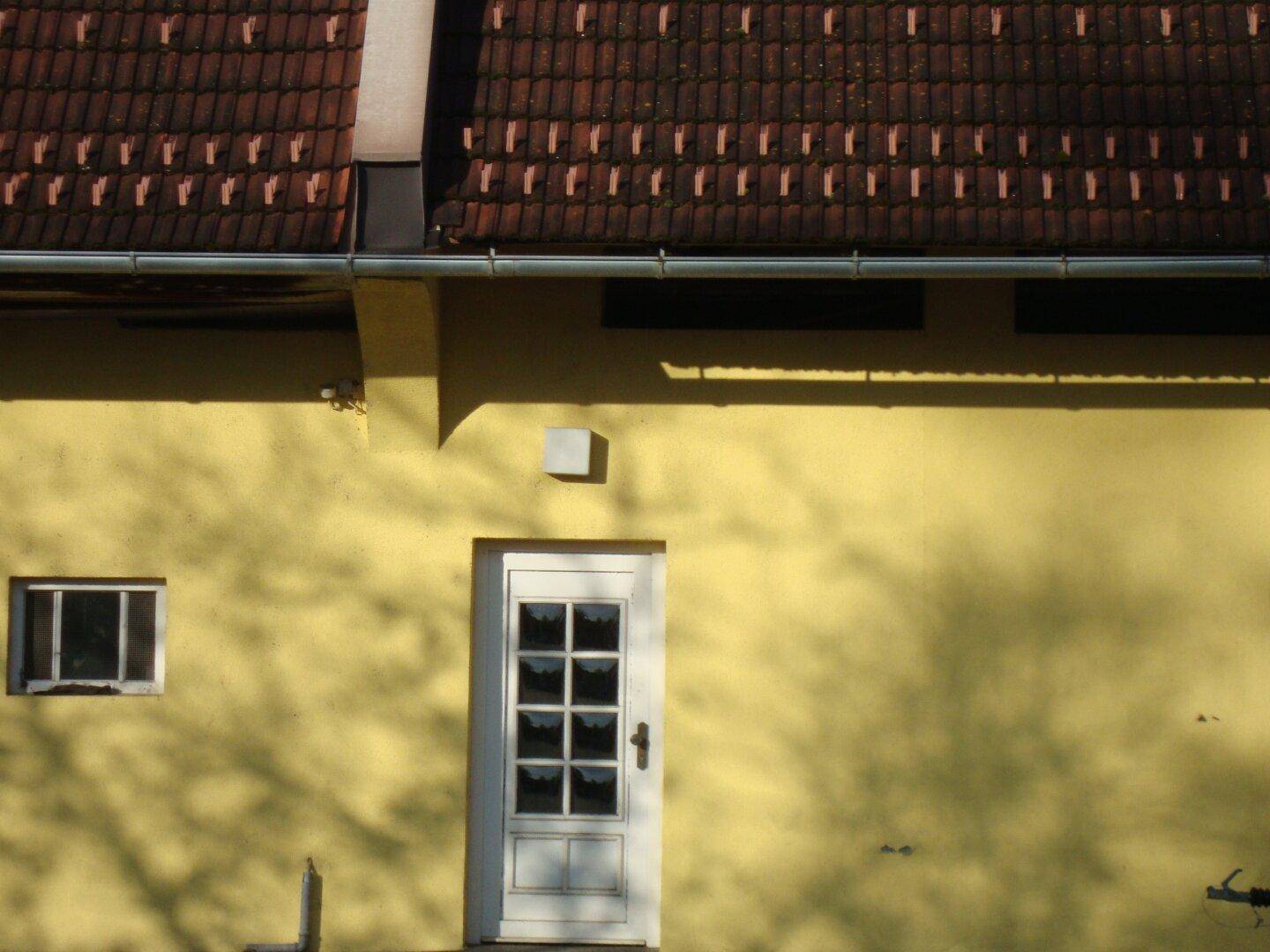 Detailansicht der gelben Fassade mit einer weißen Tür mit Glaseinsätzen und einem kleinen Fenster unter dem Ziegeldach.