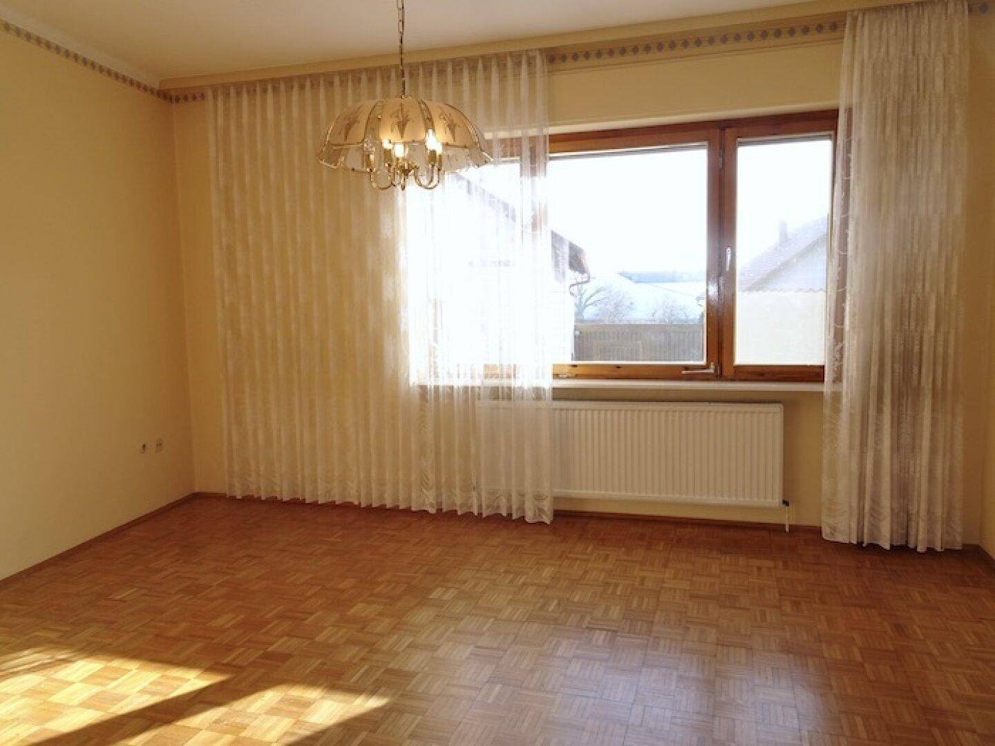 Helles Zimmer mit großem Fenster, Parkettboden und einem traditionellen Kronleuchter an der Decke.