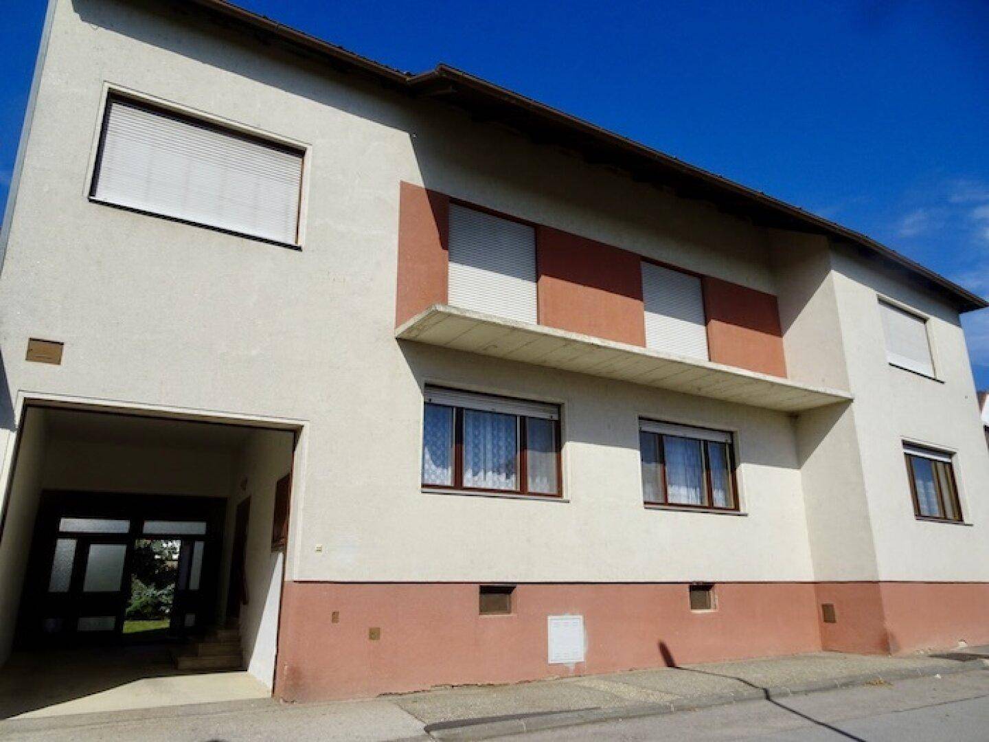 Vorderansicht des Gebäudes mit Eingangsbereich und Fenstern mit Rollläden unter klarem Himmel.