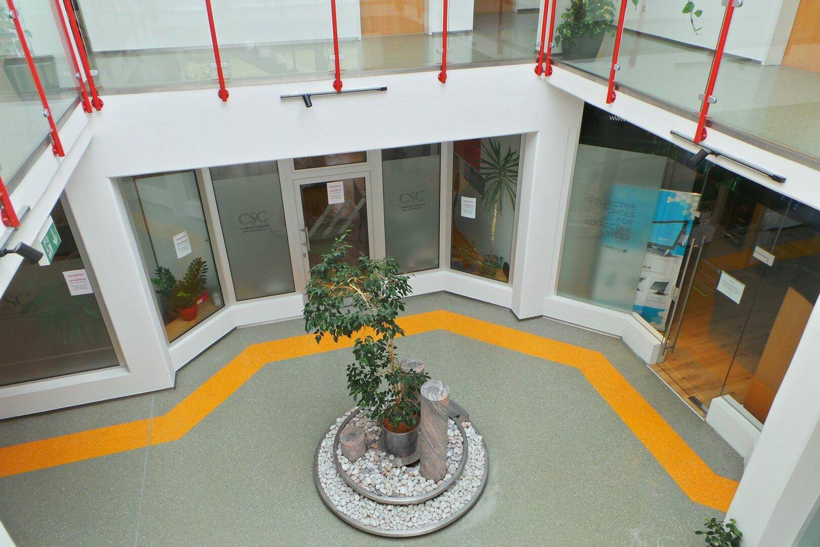 Helles Atrium mit zentraler Pflanzeninstallation, Glasgeländern und Geschäften im Erdgeschoss.