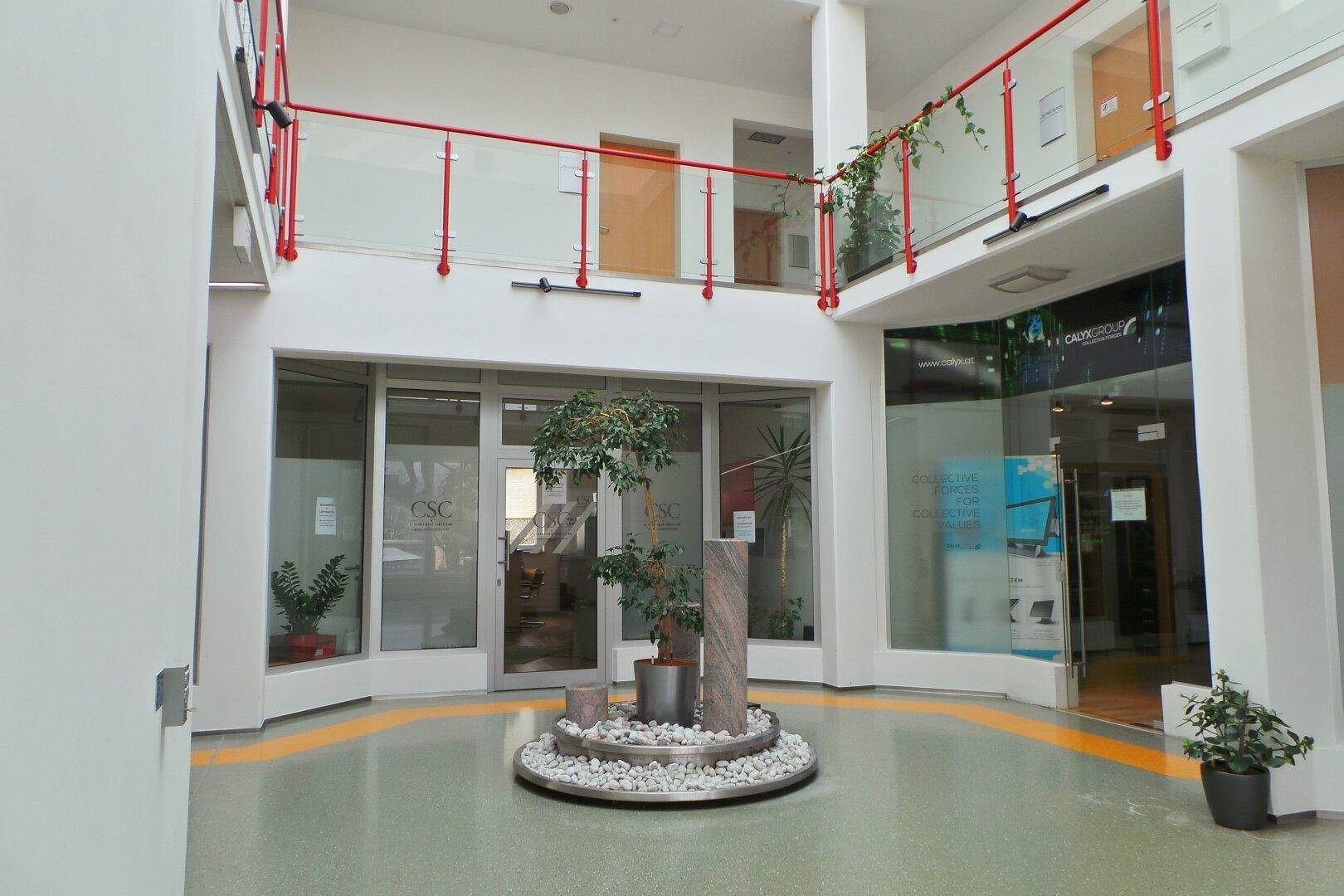 Helles Atrium mit zentraler Pflanzeninstallation, Glasgeländern und Zugang zu oberen Etagen.