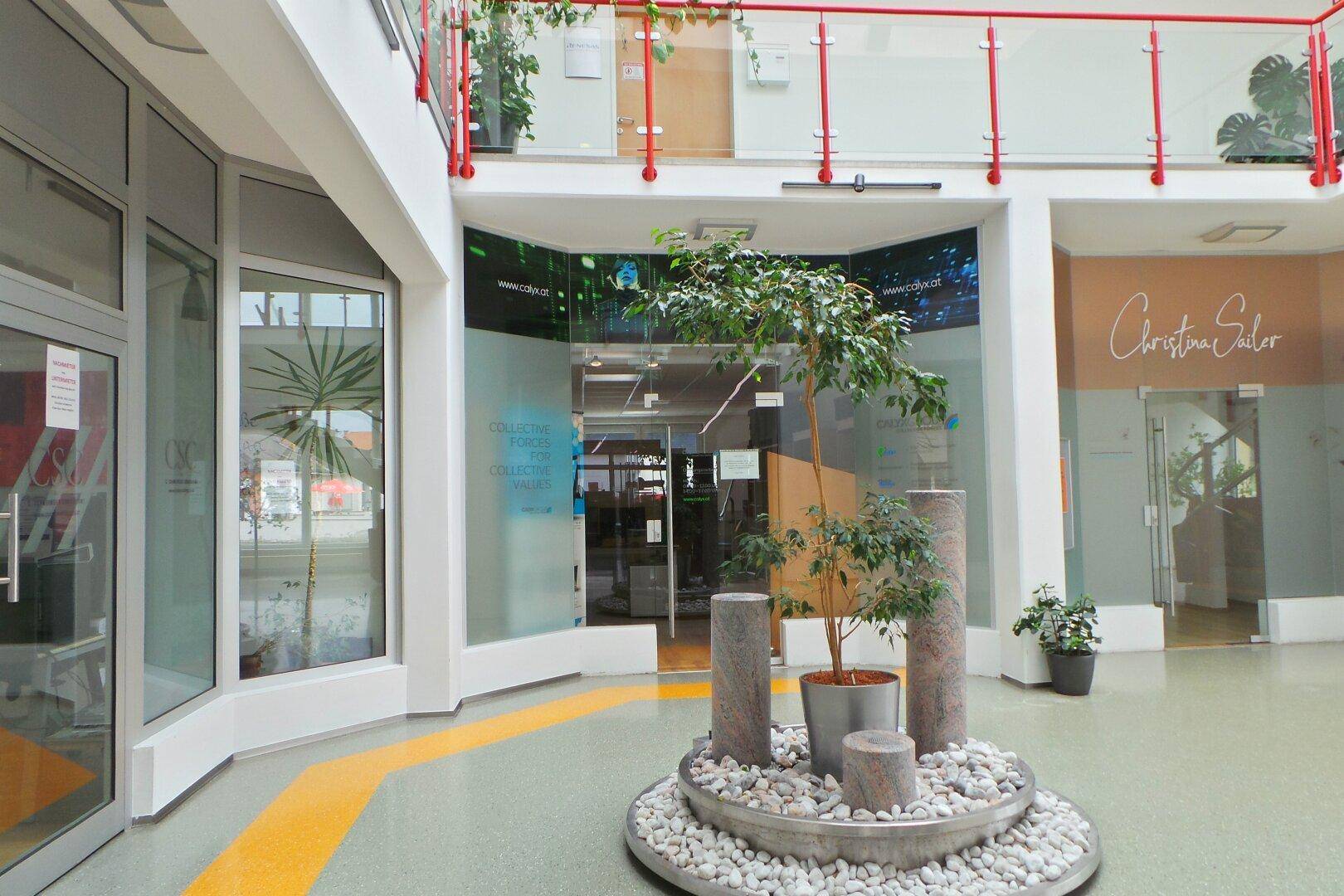 Modernes Atrium mit dekorativer Pflanzeninstallation, Glasfronten und Zugang zu verschiedenen Geschäften.