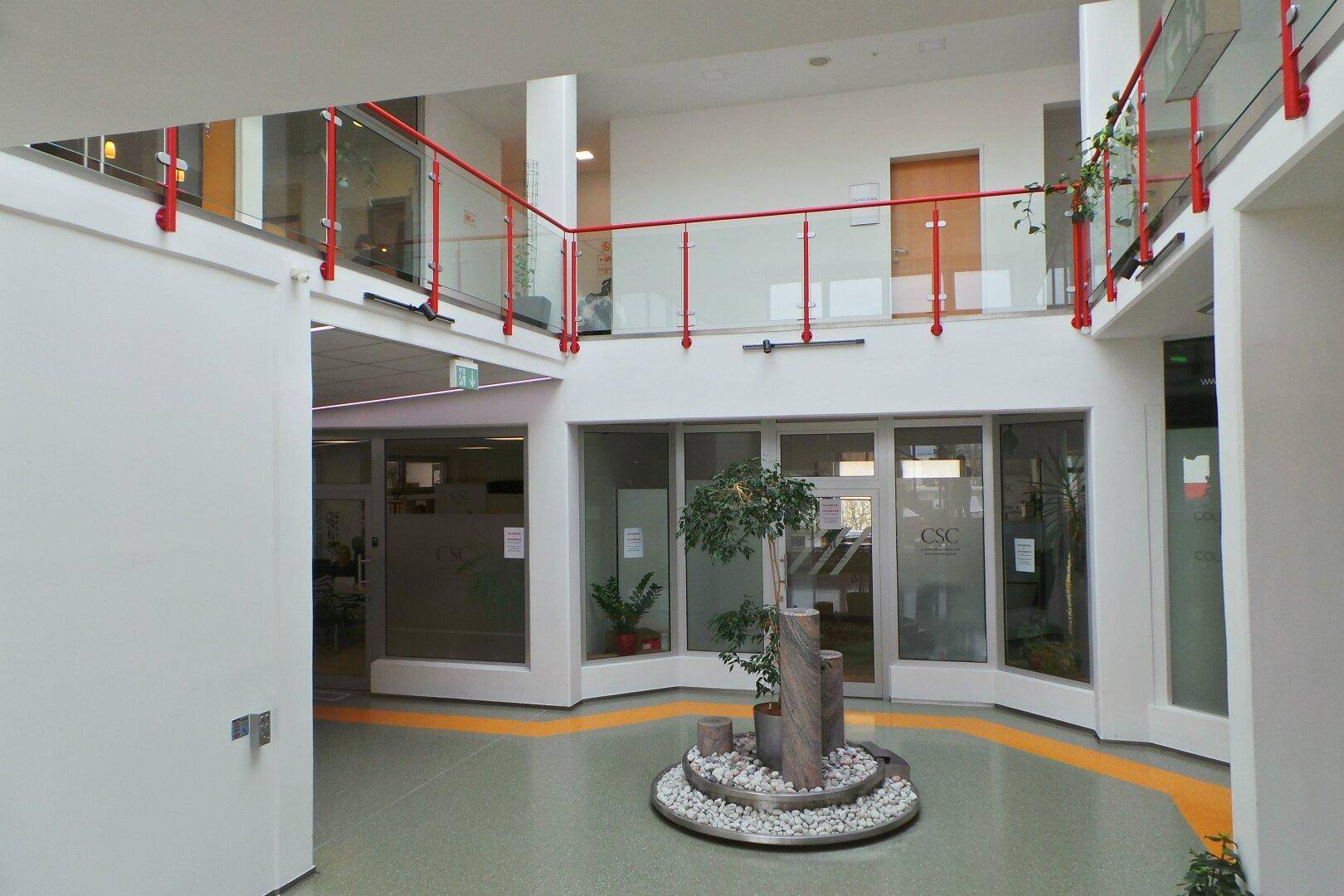 Geräumige, helle Lobby mit Glasgeländern, roten Akzenten und einem dekorativen Pflanzenarrangement.