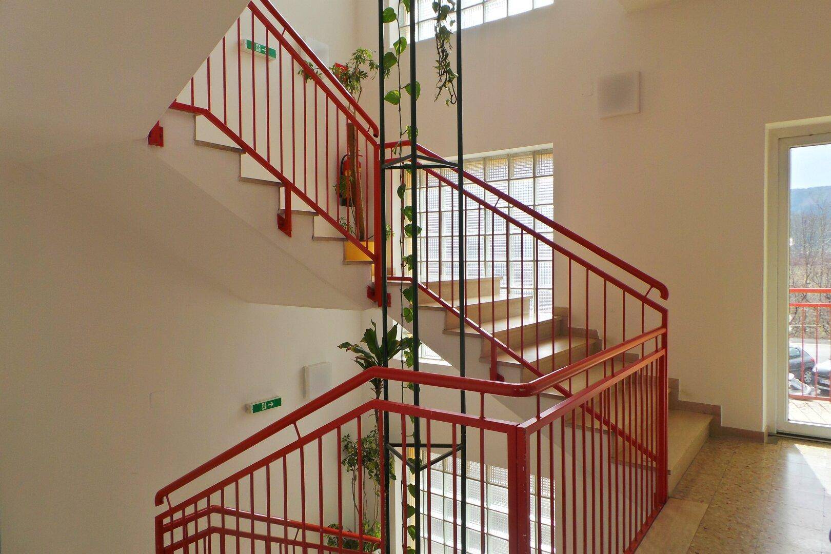 Helles Treppenhaus mit roten Geländern, großen Glasbausteinen und dekorativen Pflanzen.