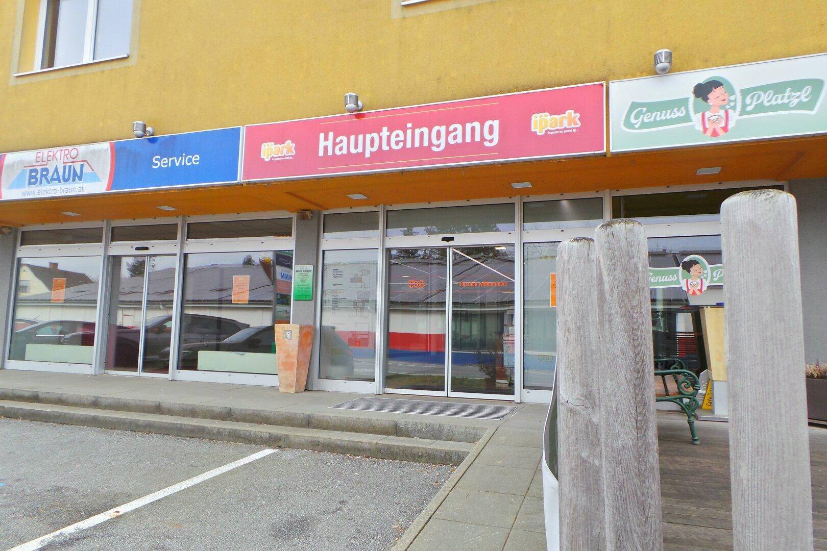 Haupteingang eines Geschäftsgebäudes mit mehreren Ladenfronten und automatischen Glastüren.