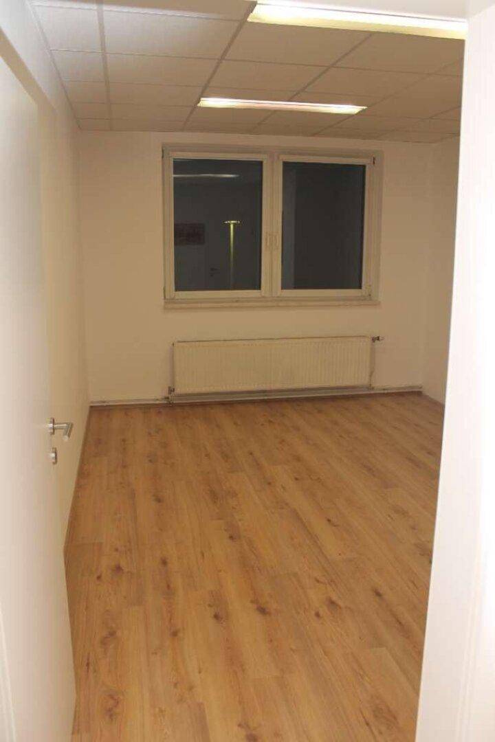 Leerer Büroraum mit Holzboden, weißen Wänden und einem Fenster mit Heizkörper darunter.