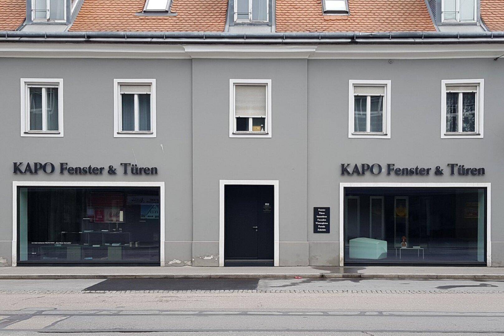 Graue Geschäftsfassade mit großen Schaufenstern und dem Schriftzug 'KAPO Fenster & Türen', darüber ein rotes Ziegeldach.