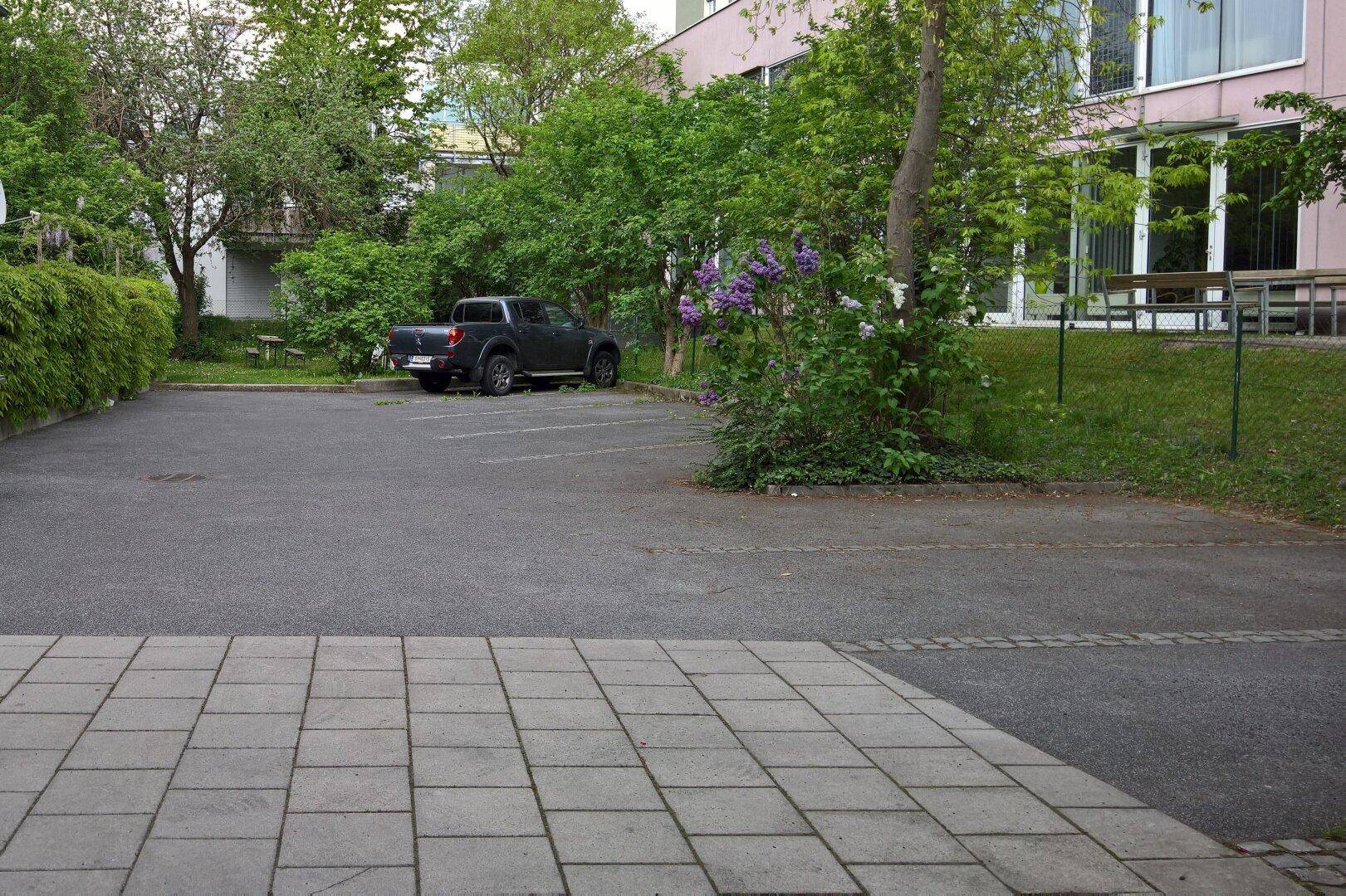 Großer asphaltierter Parkplatz mit einigen Bäumen und Sträuchern, angrenzend an ein Gebäude mit großen Fenstern.