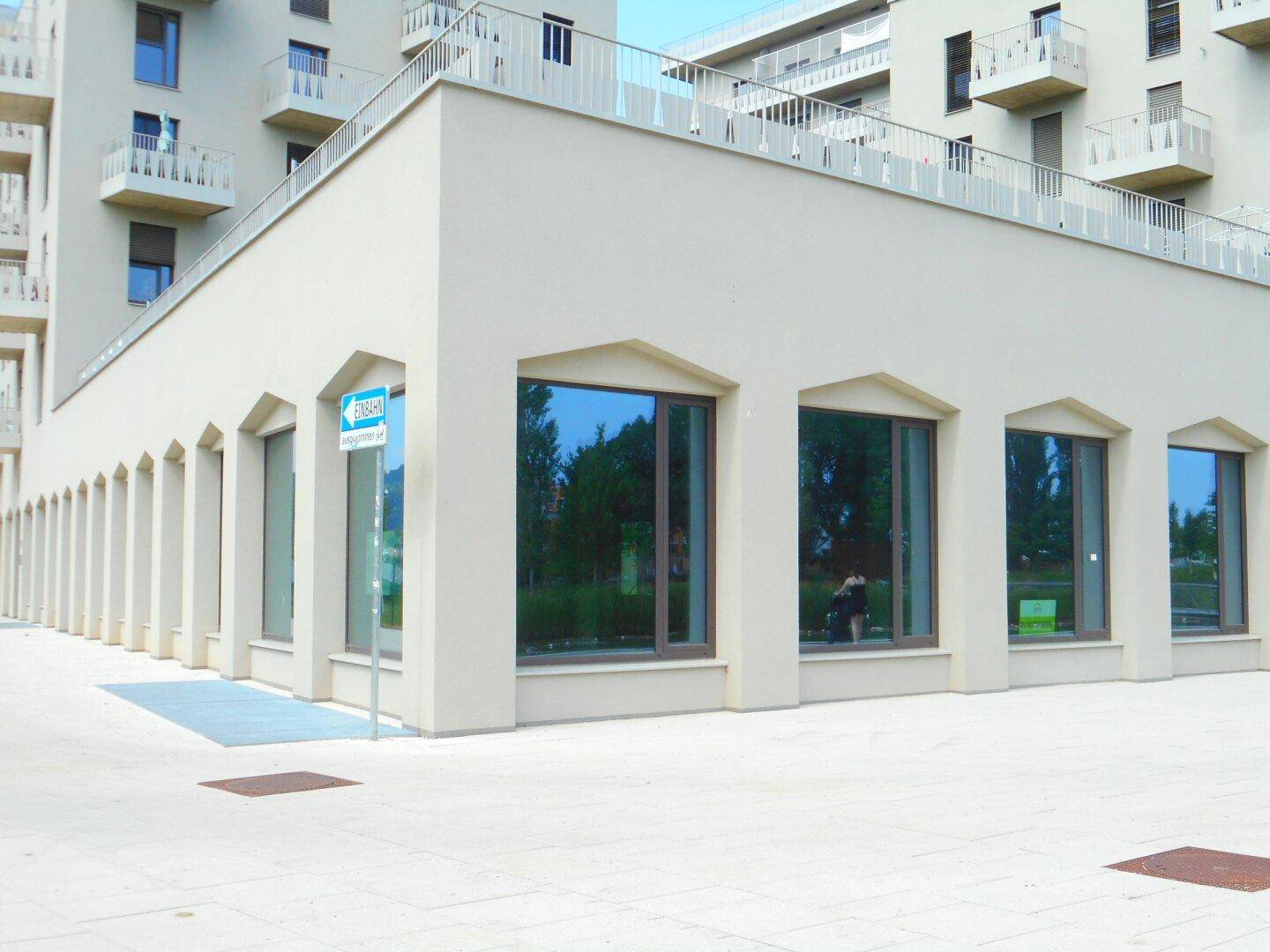 Eckansicht des modernen Gebäudes mit großen Fenstern und einer gepflasterten Fläche davor, umgeben von weiteren Neubauten.