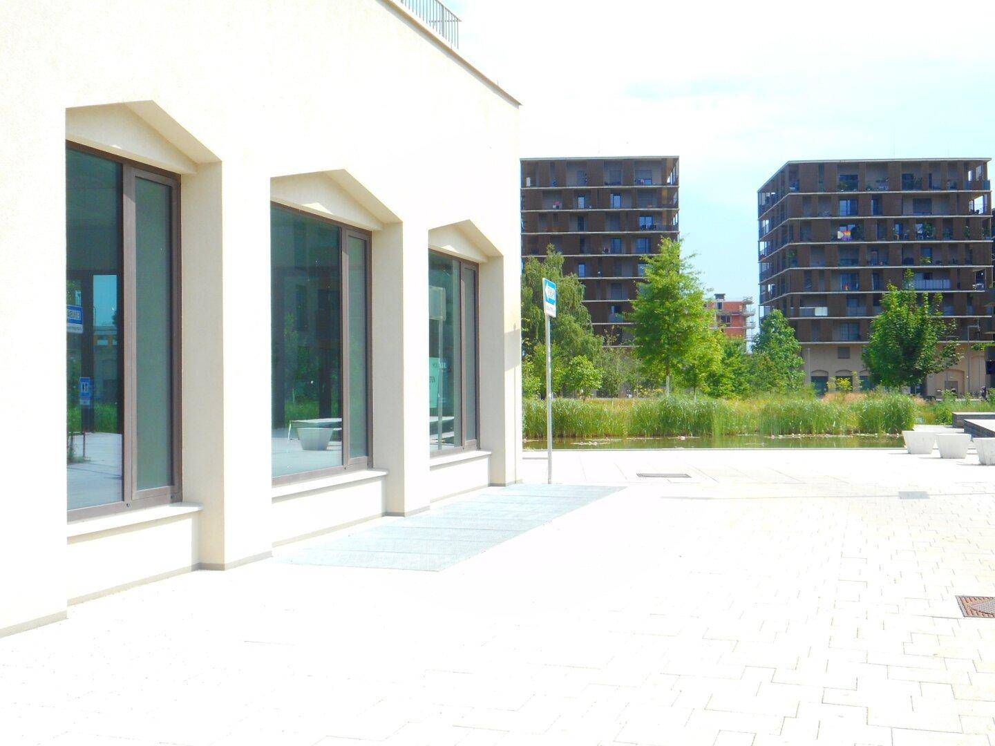Nahaufnahme der Gebäudefassade mit großen Fenstern und Blick auf eine Grünfläche mit weiteren modernen Gebäuden im Hintergrund.