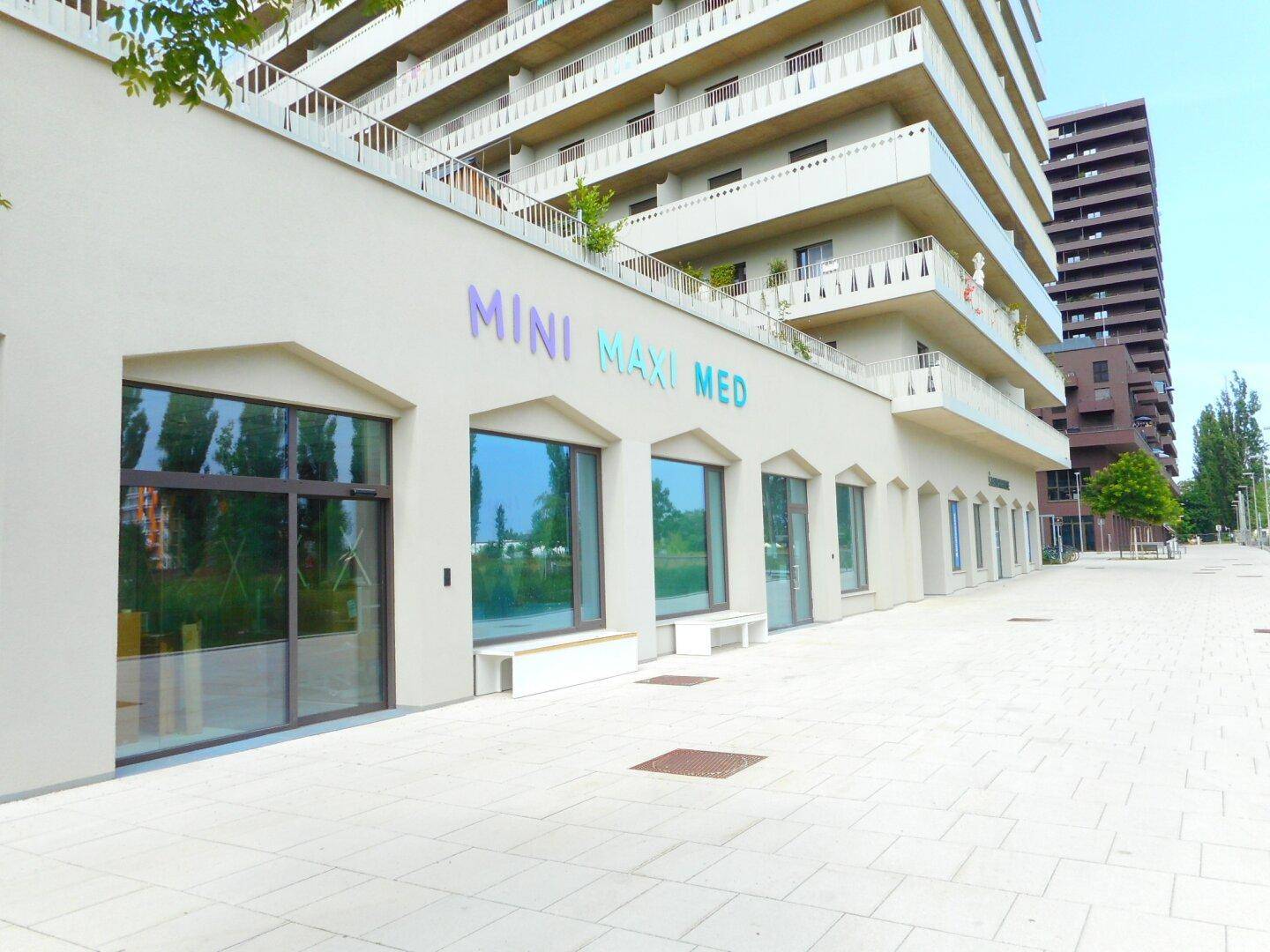 Moderne Geschäftsfassade mit großen Schaufenstern und dem Schriftzug 'MINI MAXI MED' in einem neuen Gebäudekomplex.