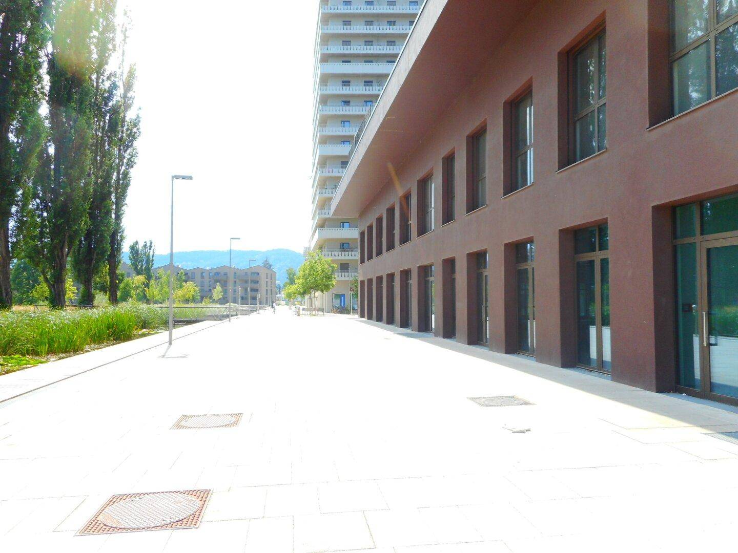 Moderne Gebäudefassade mit großen Fenstern und einem breiten, gepflasterten Gehweg im Sonnenlicht.