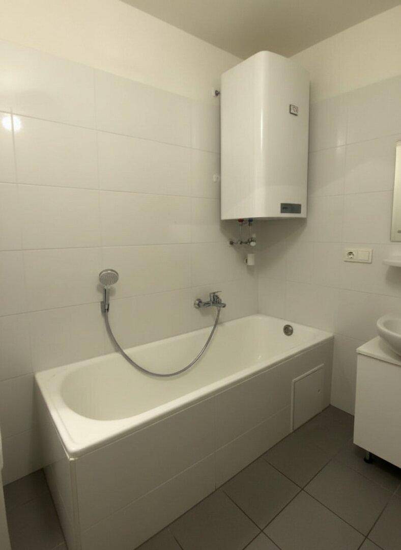 Badezimmer mit Badewanne, weißen Wandfliesen und einem Warmwasserboiler an der Wand.