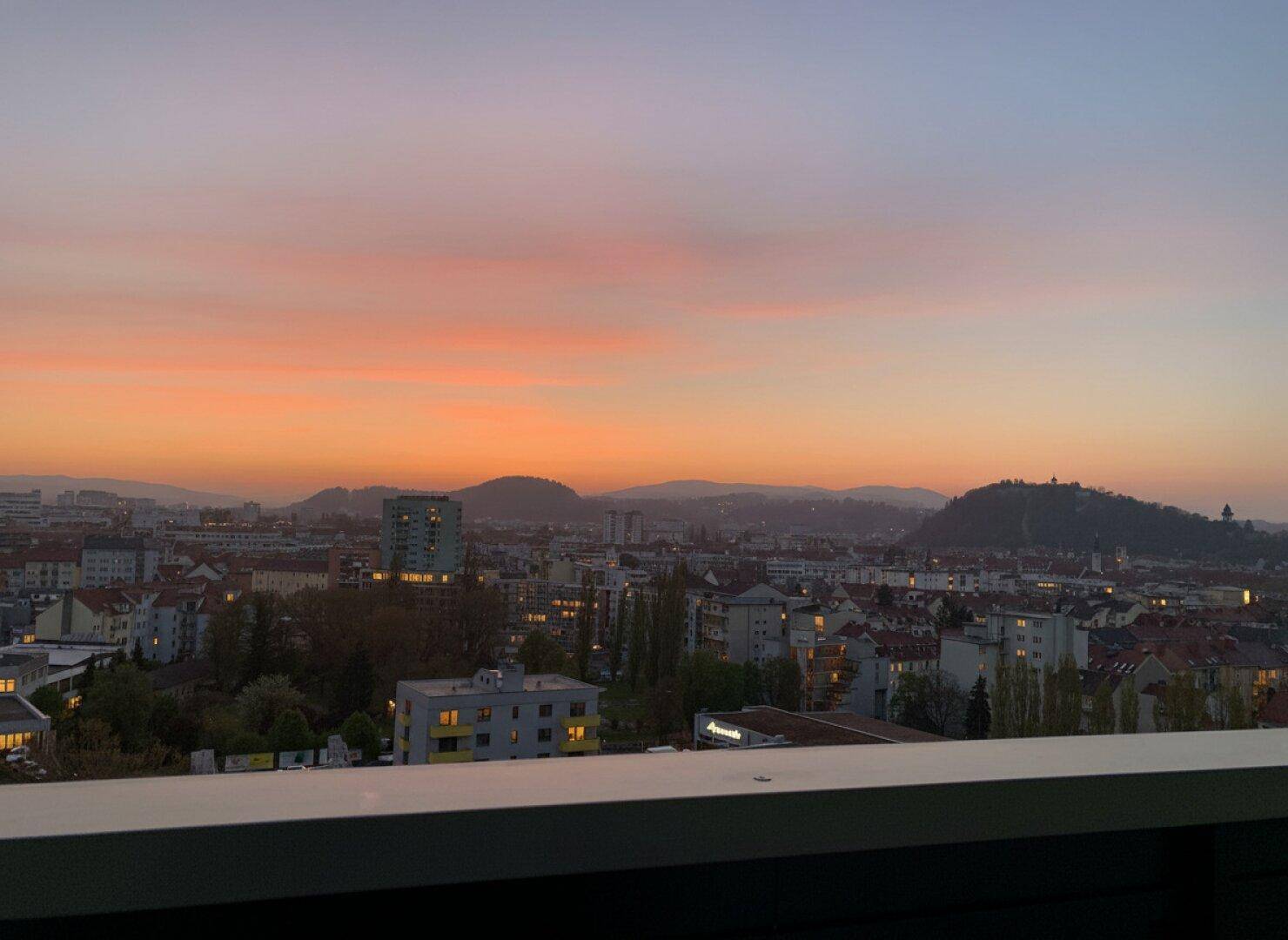 Malerischer Sonnenuntergang über der Stadt mit leuchtendem Himmel, Gebäuden und sanften Hügeln im Hintergrund.