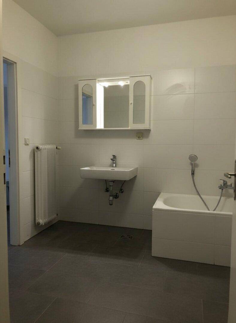 Helles Badezimmer mit Waschbecken, Spiegelschrank, Badewanne und Heizkörper, ausgestattet mit grauen Fliesen.