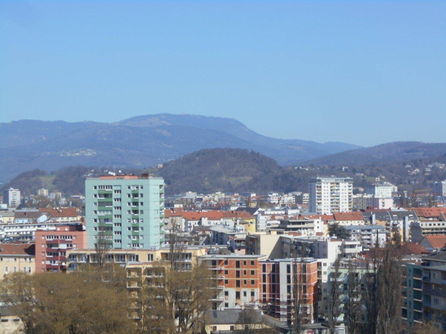 Weitläufiger Blick über die Stadtlandschaft mit modernen Gebäuden und imposanten Bergen am Horizont.