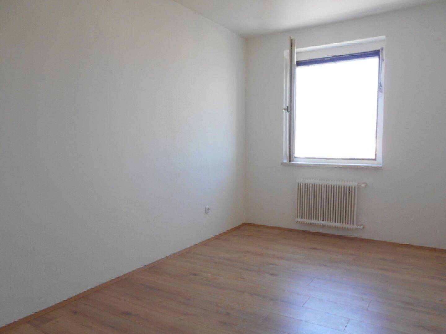 Leeres Zimmer mit Laminatboden, weiß gestrichenen Wänden und einem Fenster, bereit zur Einrichtung.