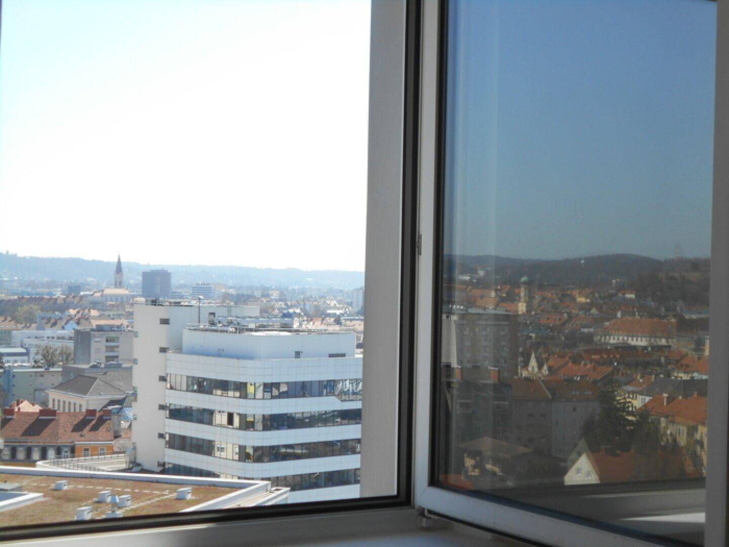 Ausblick aus dem Fenster auf die städtische Umgebung mit Gebäuden und grünen Hügeln in der Ferne.