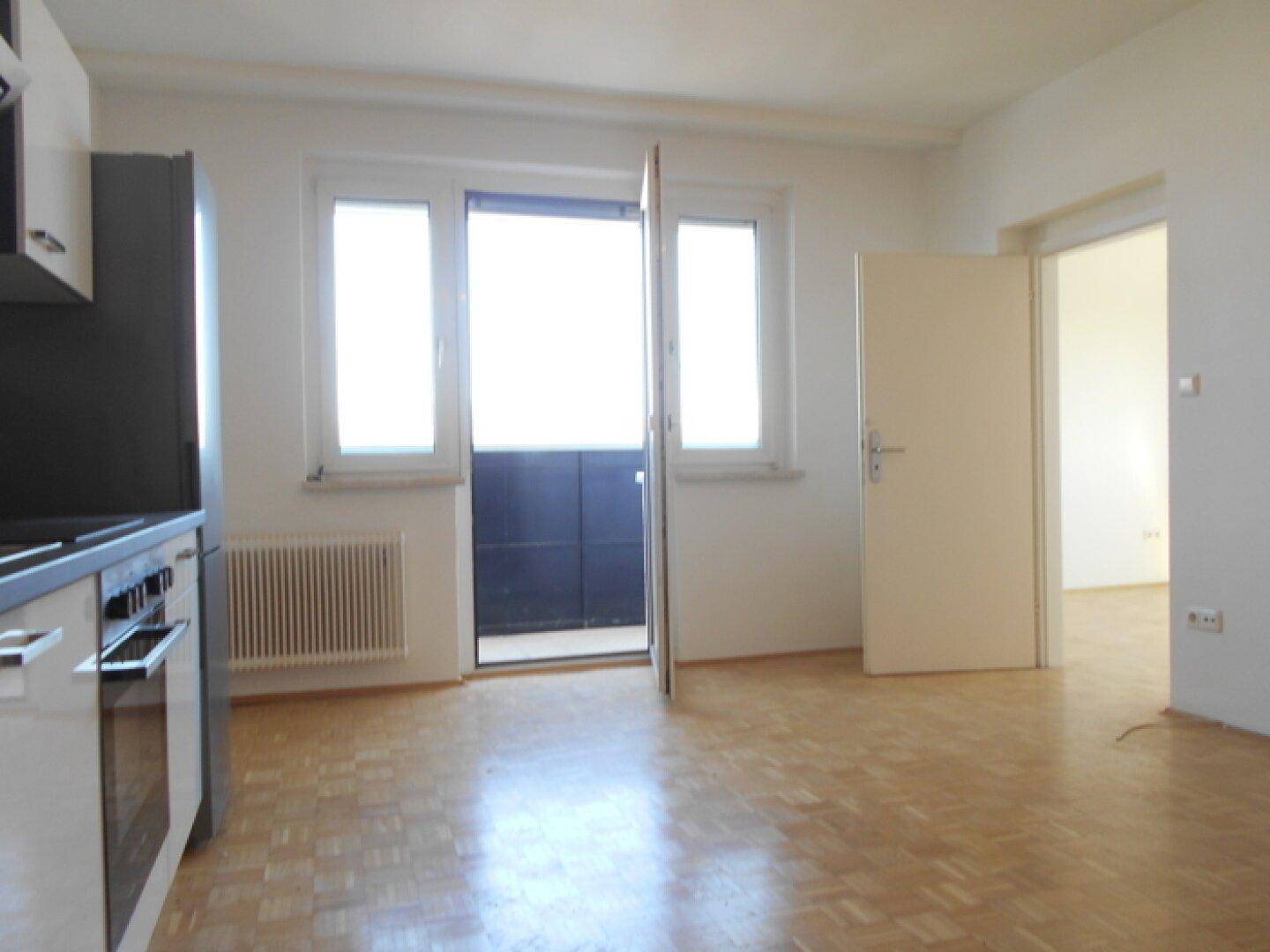 Heller Wohnbereich mit Parkettboden, Heizkörper und Zugang zu einem Balkon, verbunden mit einem weiteren Zimmer.