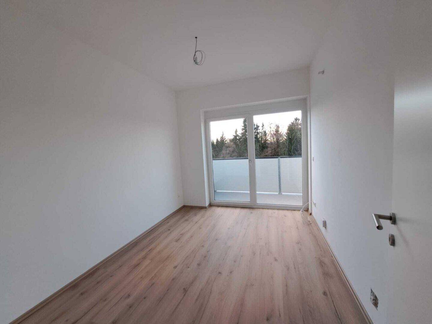 Heller, leerer Raum mit Laminatboden und großem Fenster mit Zugang zum Balkon.