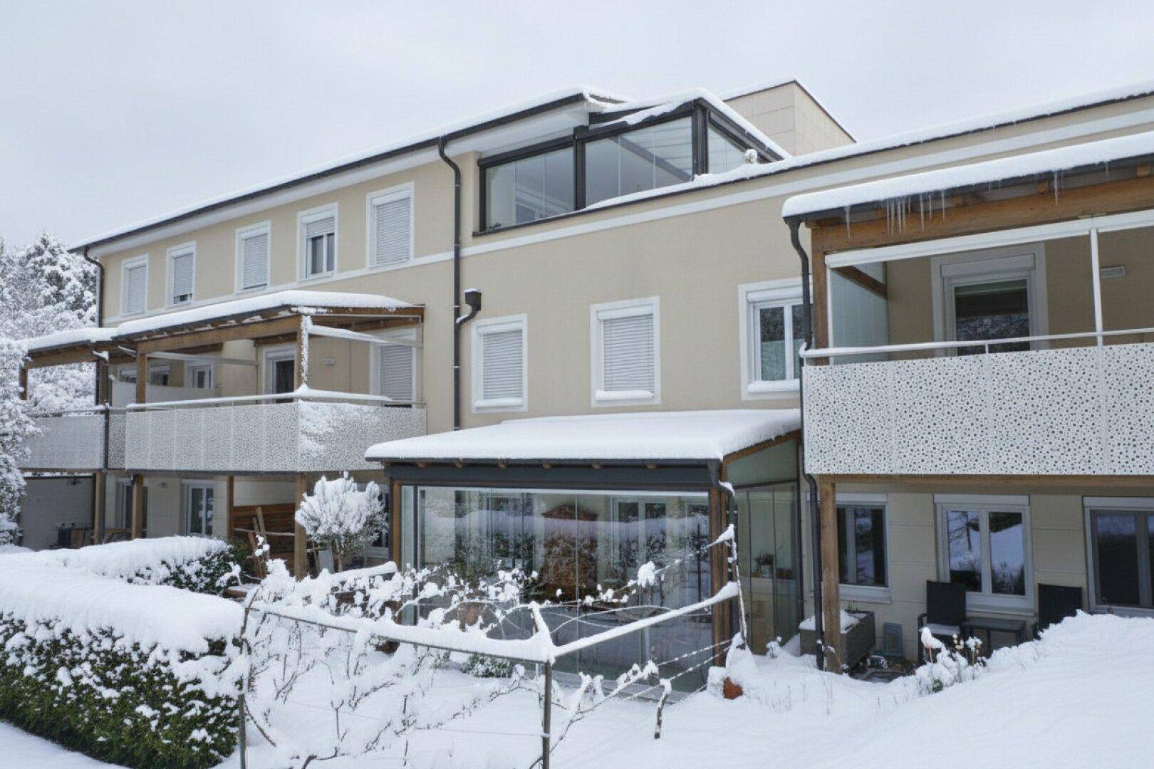 Mehrfamilienhaus im Winter mit schneebedeckten Balkonen, einem Wintergarten und einer Dachverglasung.
