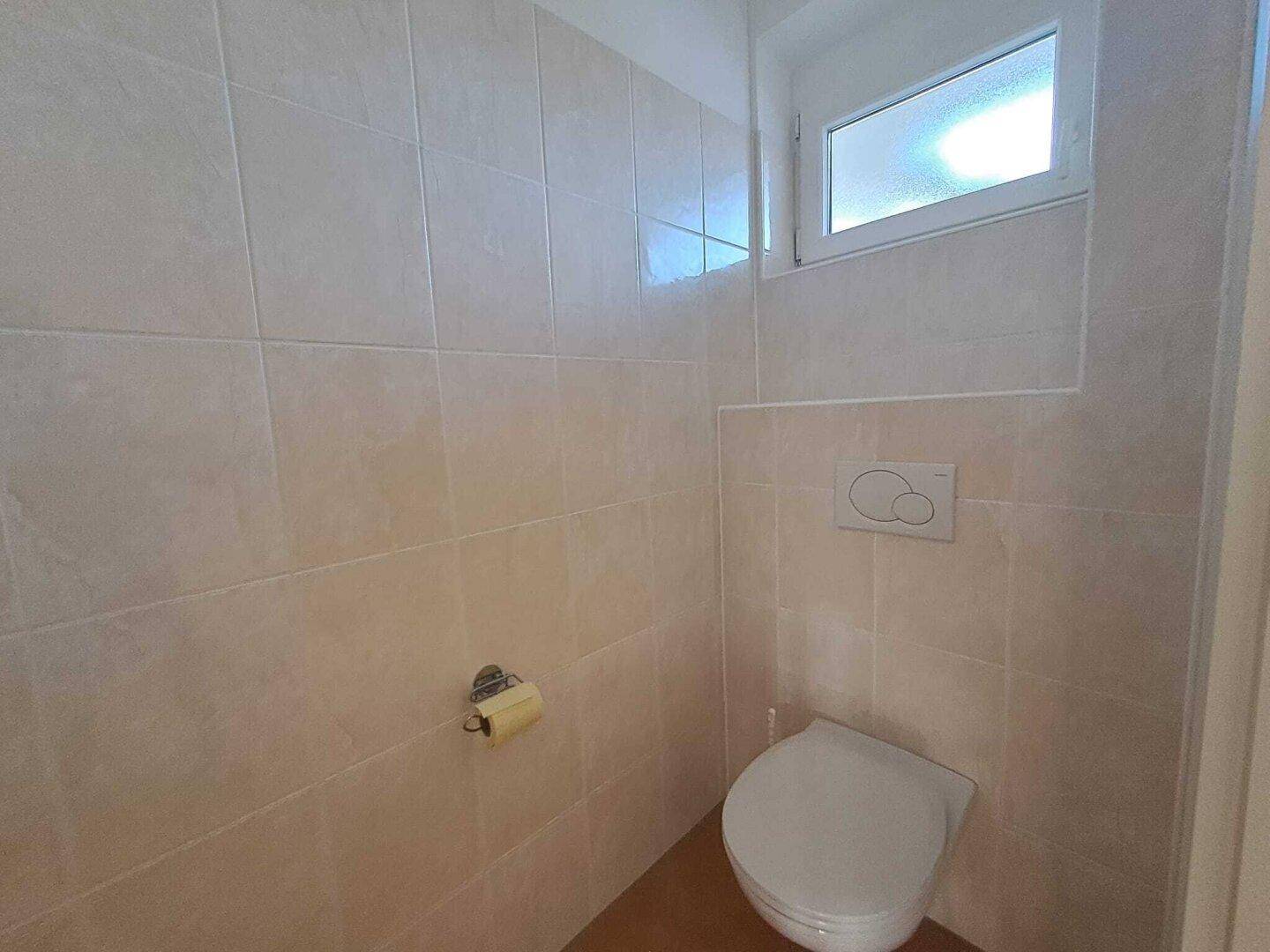 Separates WC mit wandhängender Toilette und beigefarbenen Fliesen.