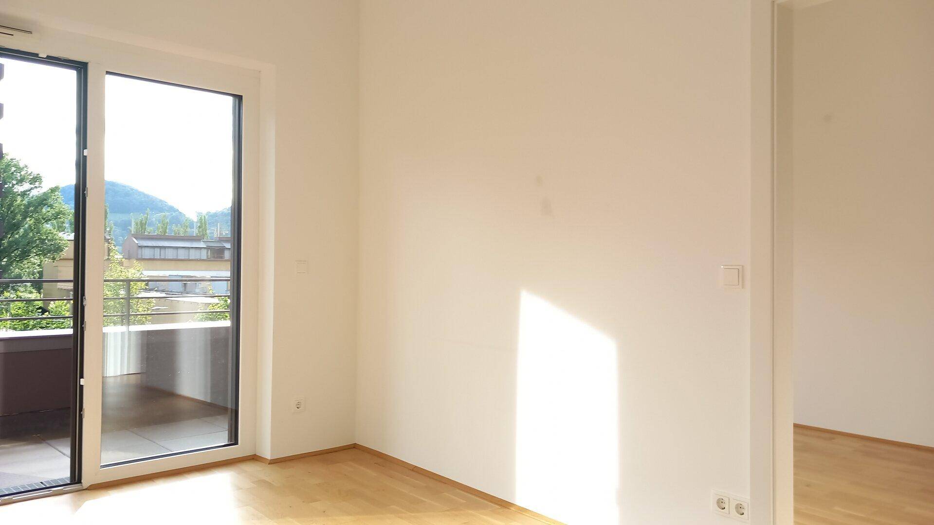 Geräumiger Wohnbereich mit Holzboden und großen Fenstern, die einen weiten Blick ins Grüne ermöglichen.