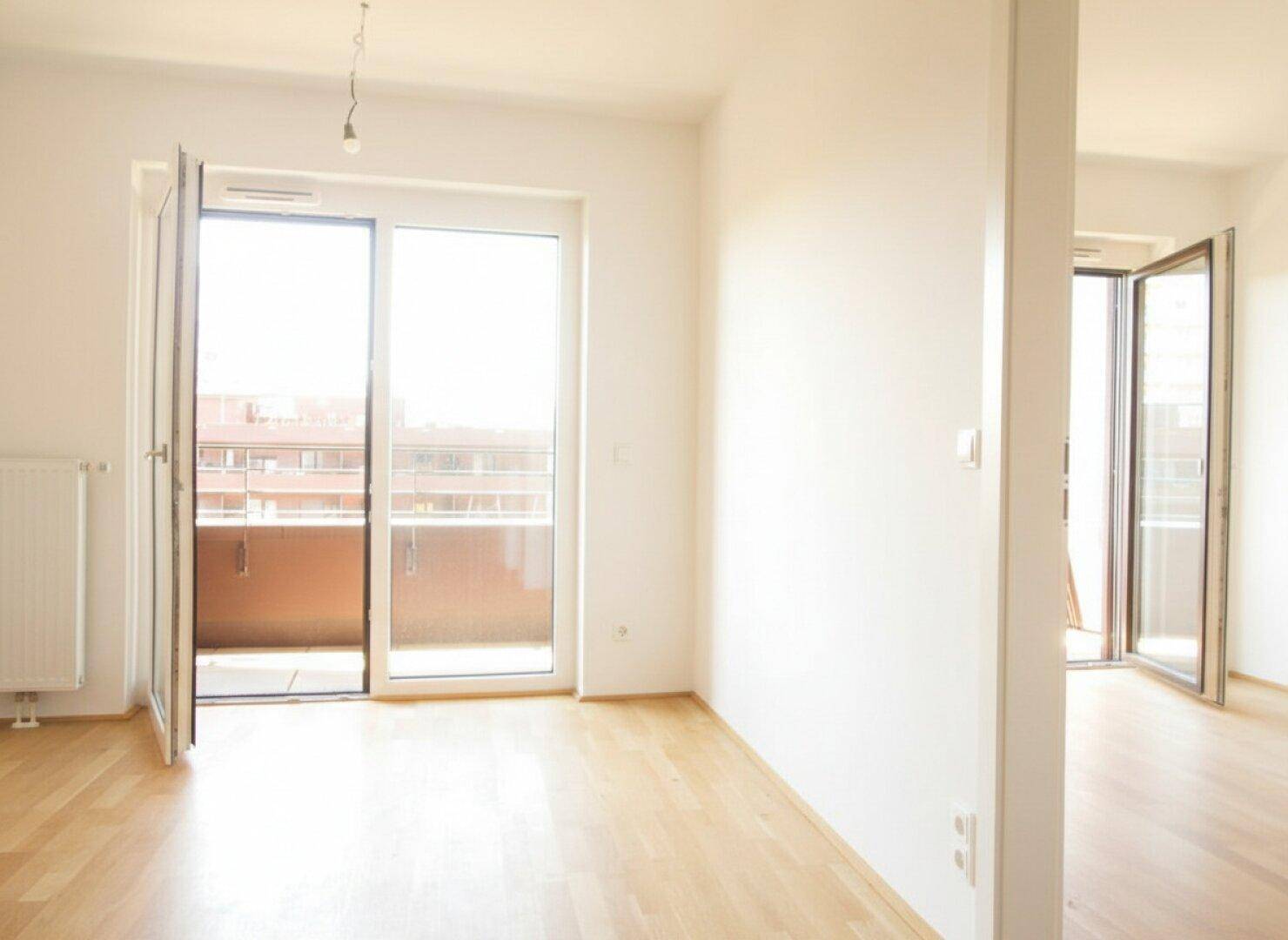 Heller Wohnraum mit Holzboden und großem Balkonzugang, der viel Tageslicht hereinlässt.