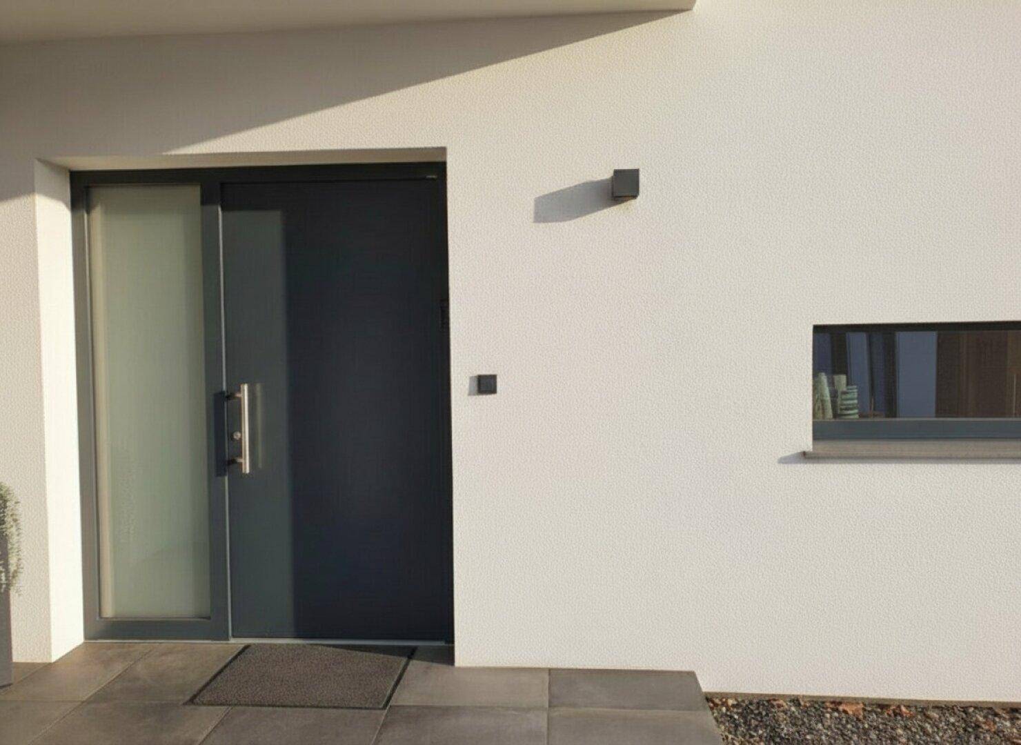 Moderner Hauseingang mit dunkler Tür und seitlichem Fenster, das für Tageslicht sorgt.