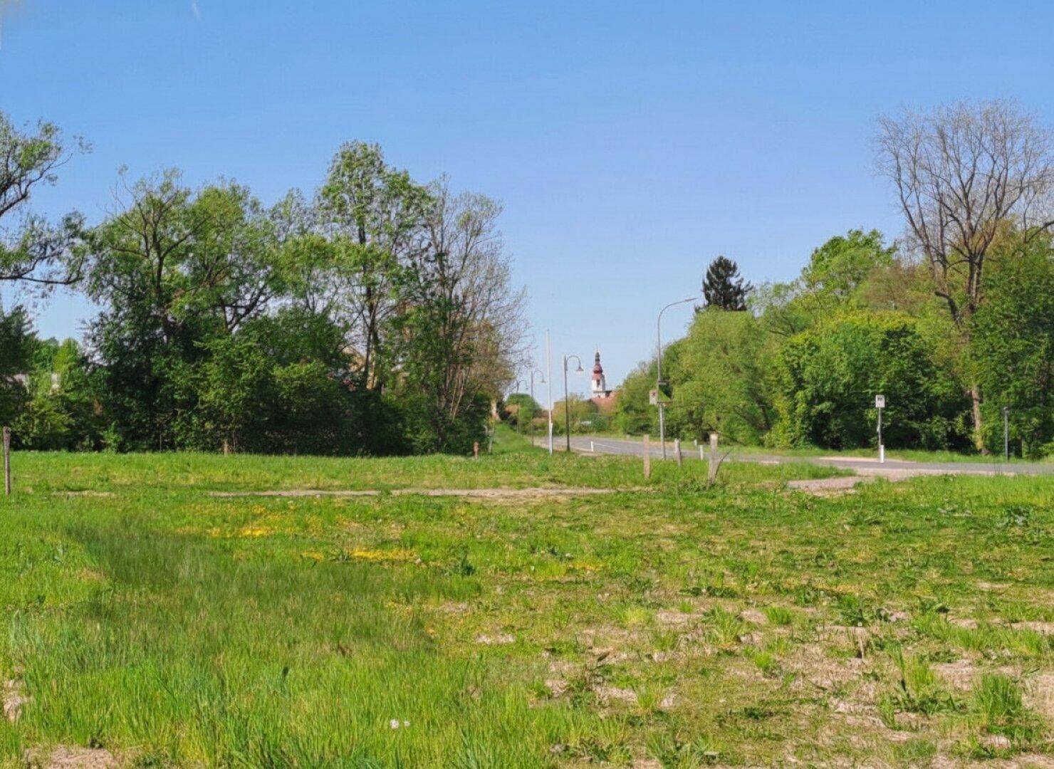 Offenes Gelände mit einer Straße, Bäumen und einem Kirchturm in der Ferne.