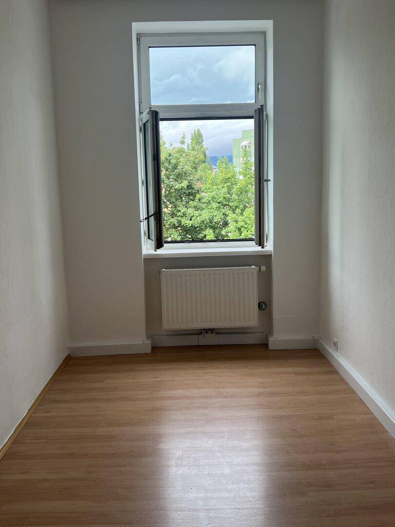 Kleiner, heller Raum mit Laminatboden und einem hohen Fenster mit Blick ins Grüne.