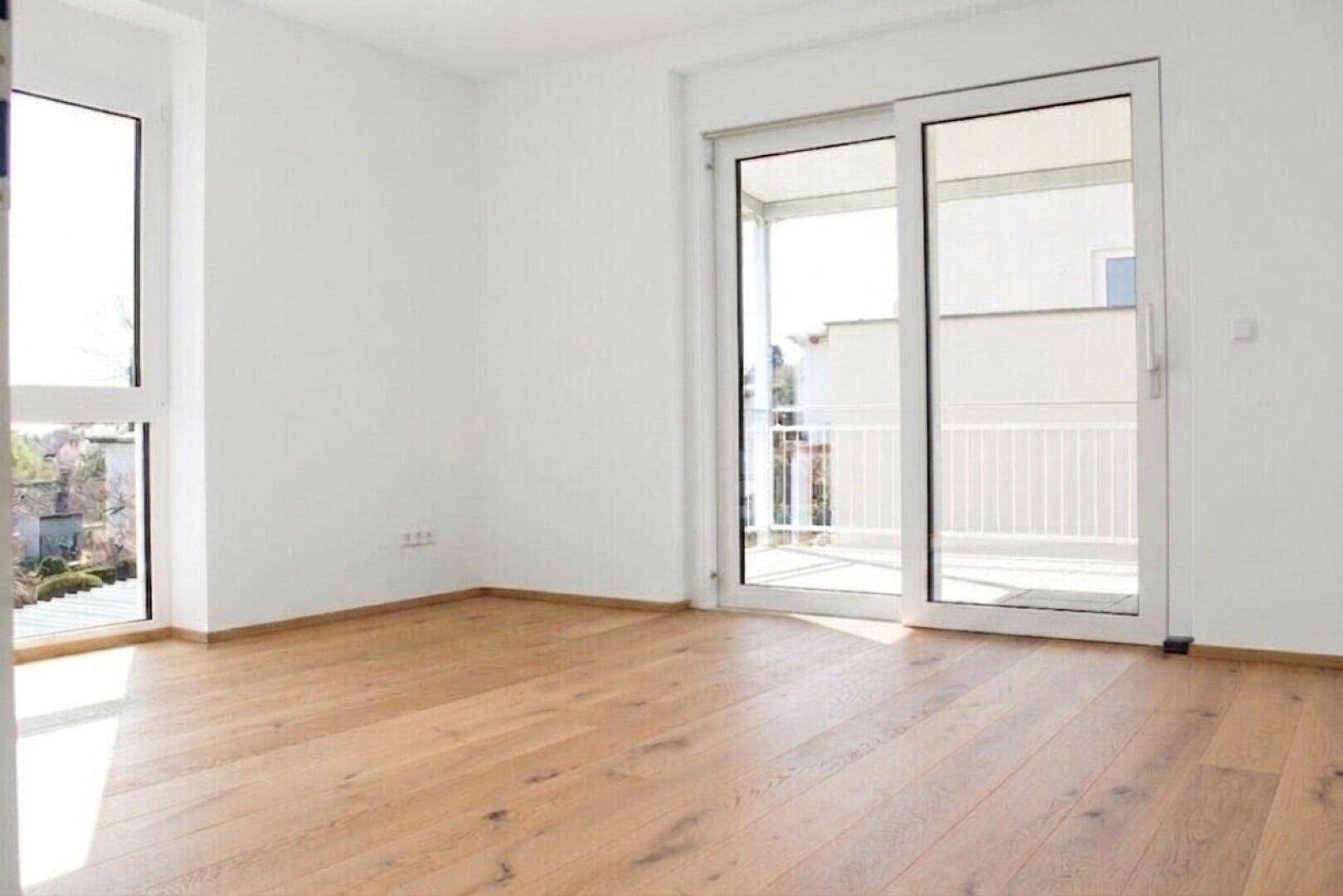 Helles leeres Wohnzimmer mit Holzboden und großem Schiebefenster zum Balkon.