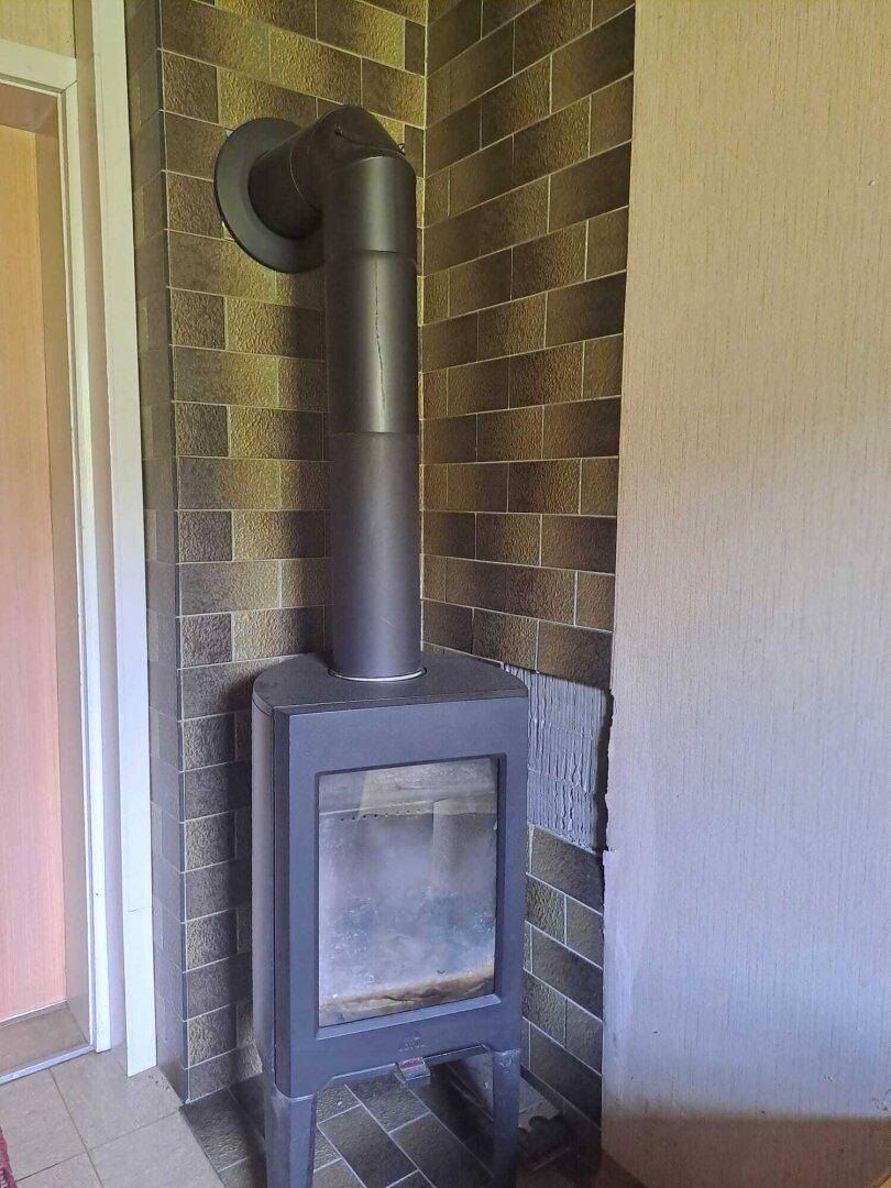 Moderner Holzofen mit Ofenrohr vor einer gefliesten Wand im Flurbereich.