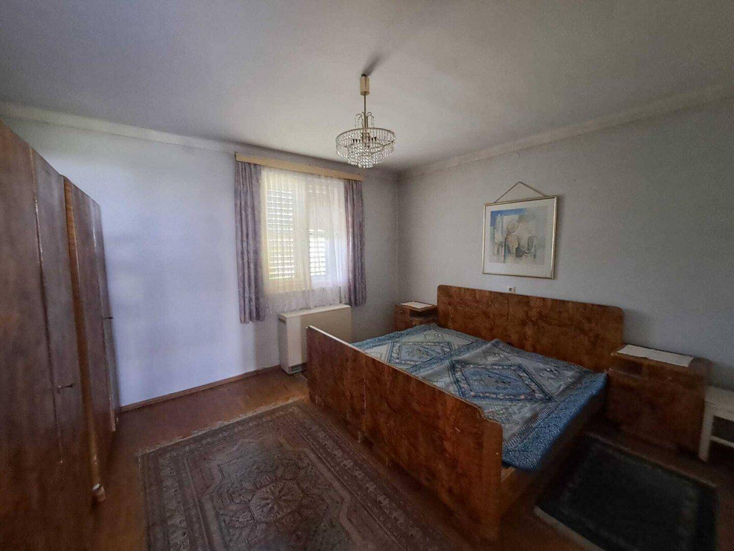 Schlafzimmer mit Holzbett, Teppich und Fenster, das viel Tageslicht hereinlässt.
