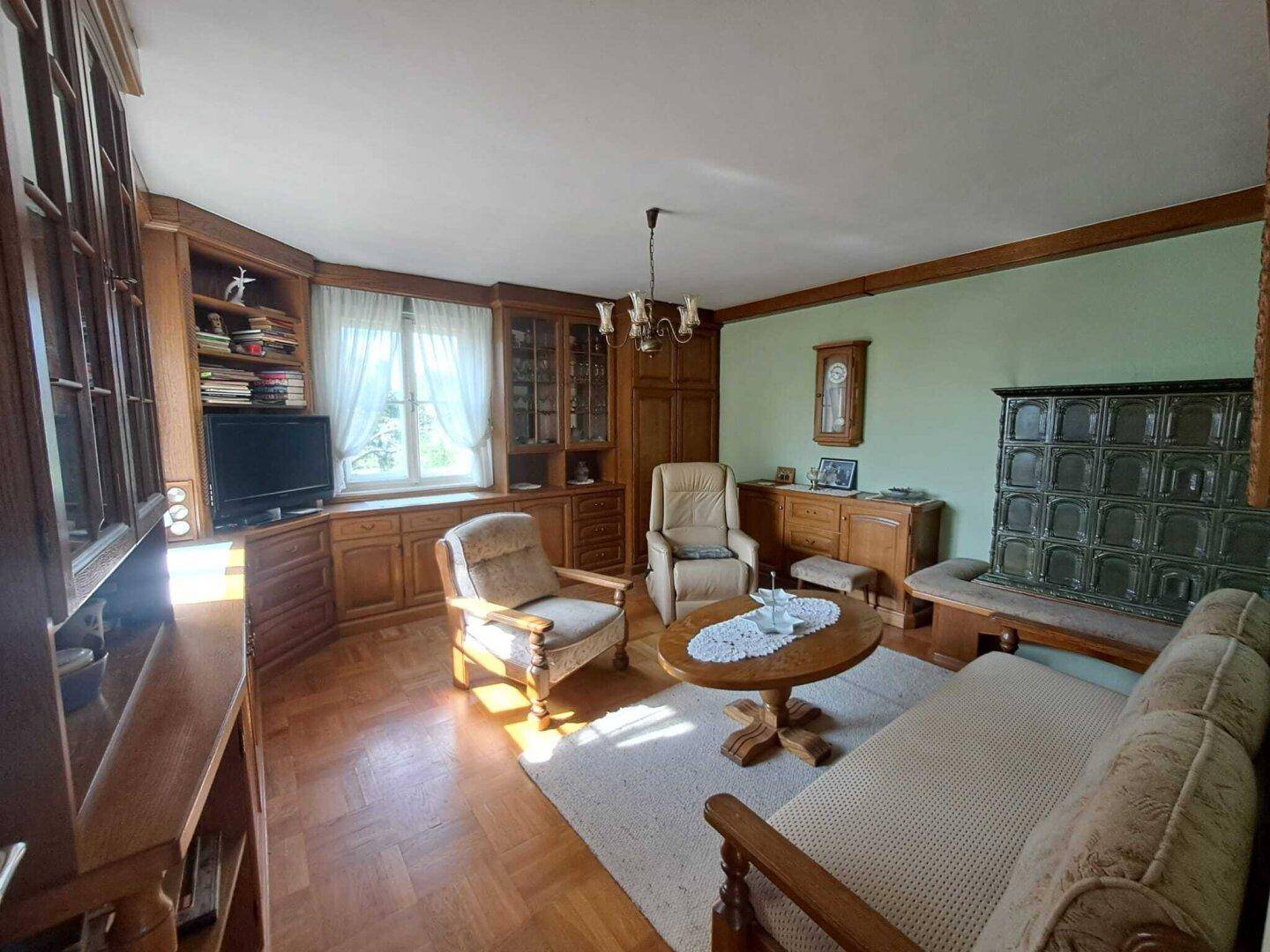 Geräumiges Wohnzimmer mit Holzboden, Kachelofen und großen Fenstern für viel Licht.