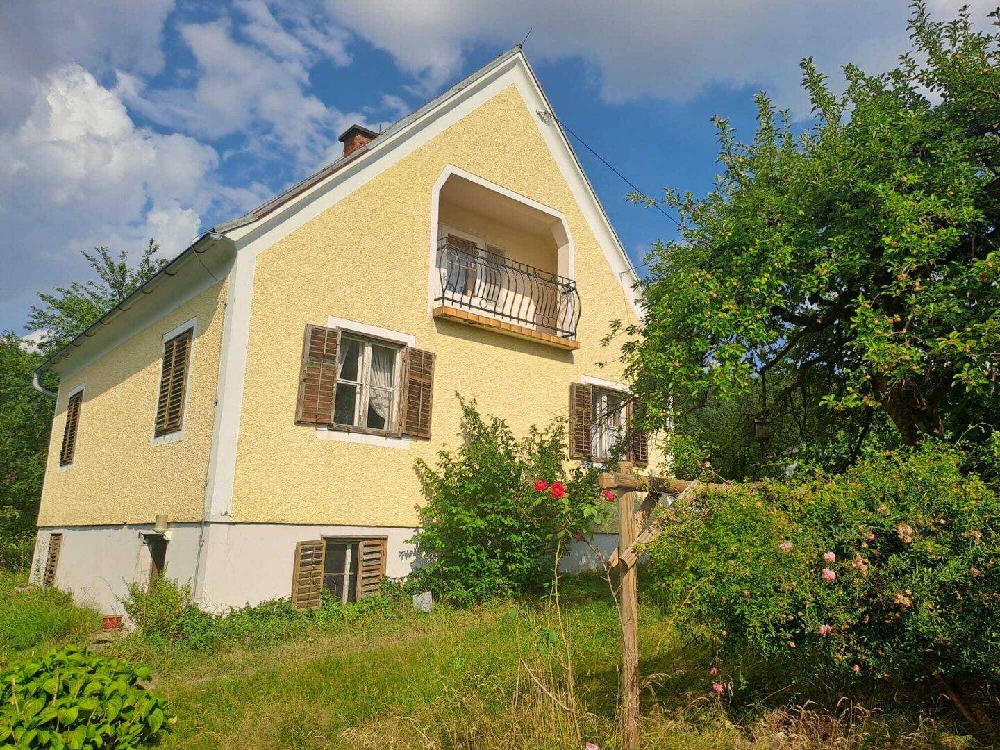 Gelbes Landhaus mit Balkon, braunen Fensterläden und umgebendem Grün.