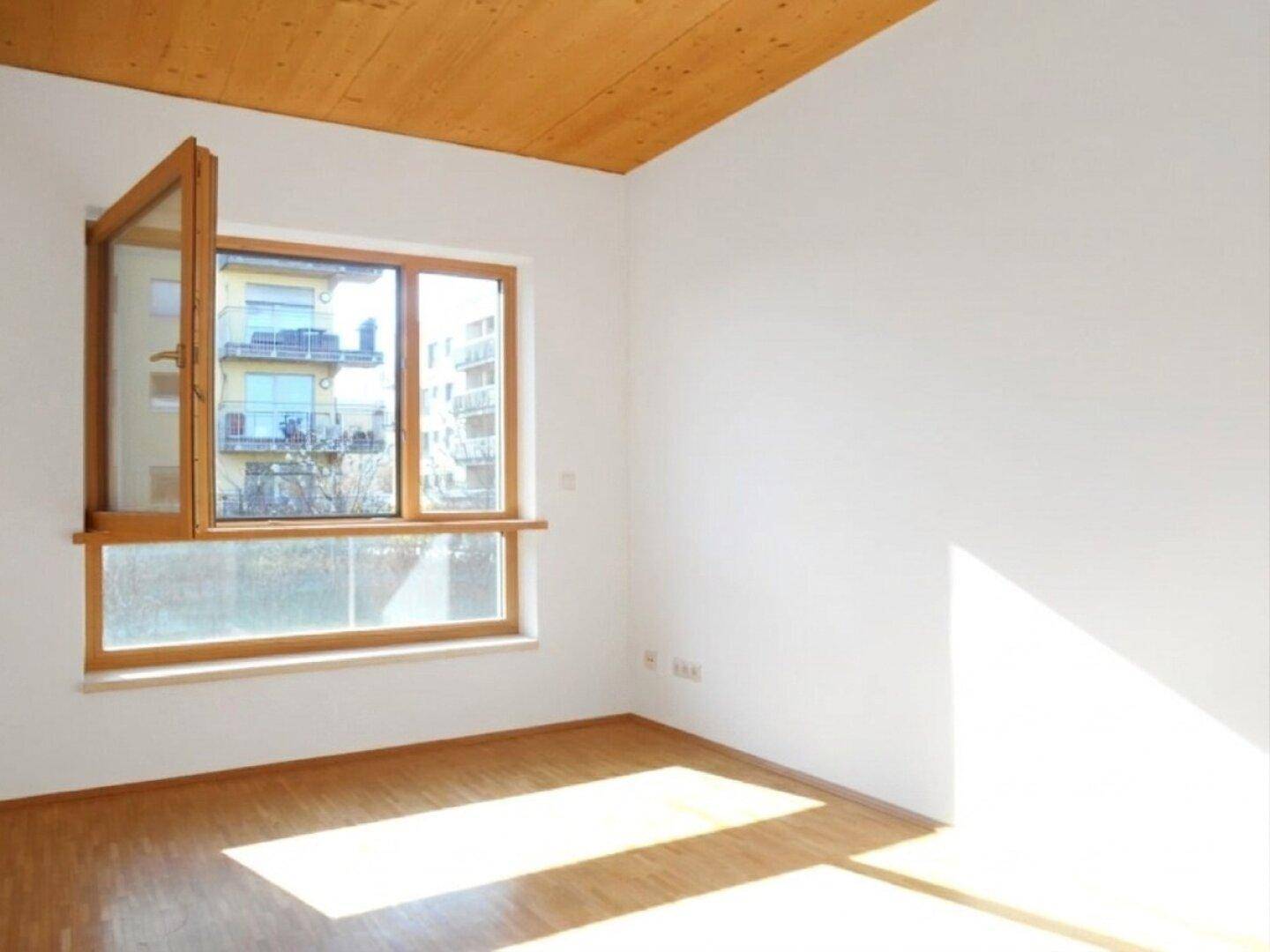 Lichtdurchflutetes Zimmer mit Holzbalkendecke, Parkettboden und einem großen Fenster mit Blick nach draußen.